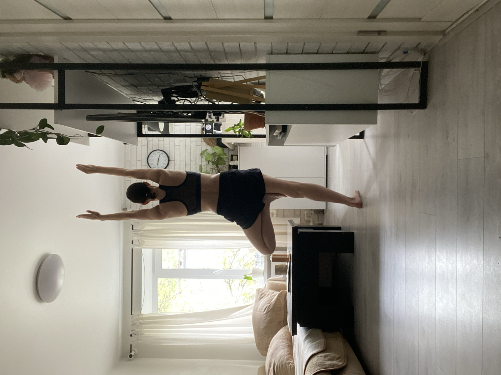 A woman is performing a yoga pose in a room. She is wearing a black sports bra and black shorts. Her right leg is extended straight to the side, and her left leg is bent, with her left foot lifted off the ground. Both arms are extended straight out, one to the right and one to the left. The room has white walls and a light wood floor. There is a black table and a light-colored couch in the background. A window with white curtains is visible in the background. The image appears to have been taken inside a home, possibly in Chaikovsky, Russia. A wall clock can be seen in the background.
