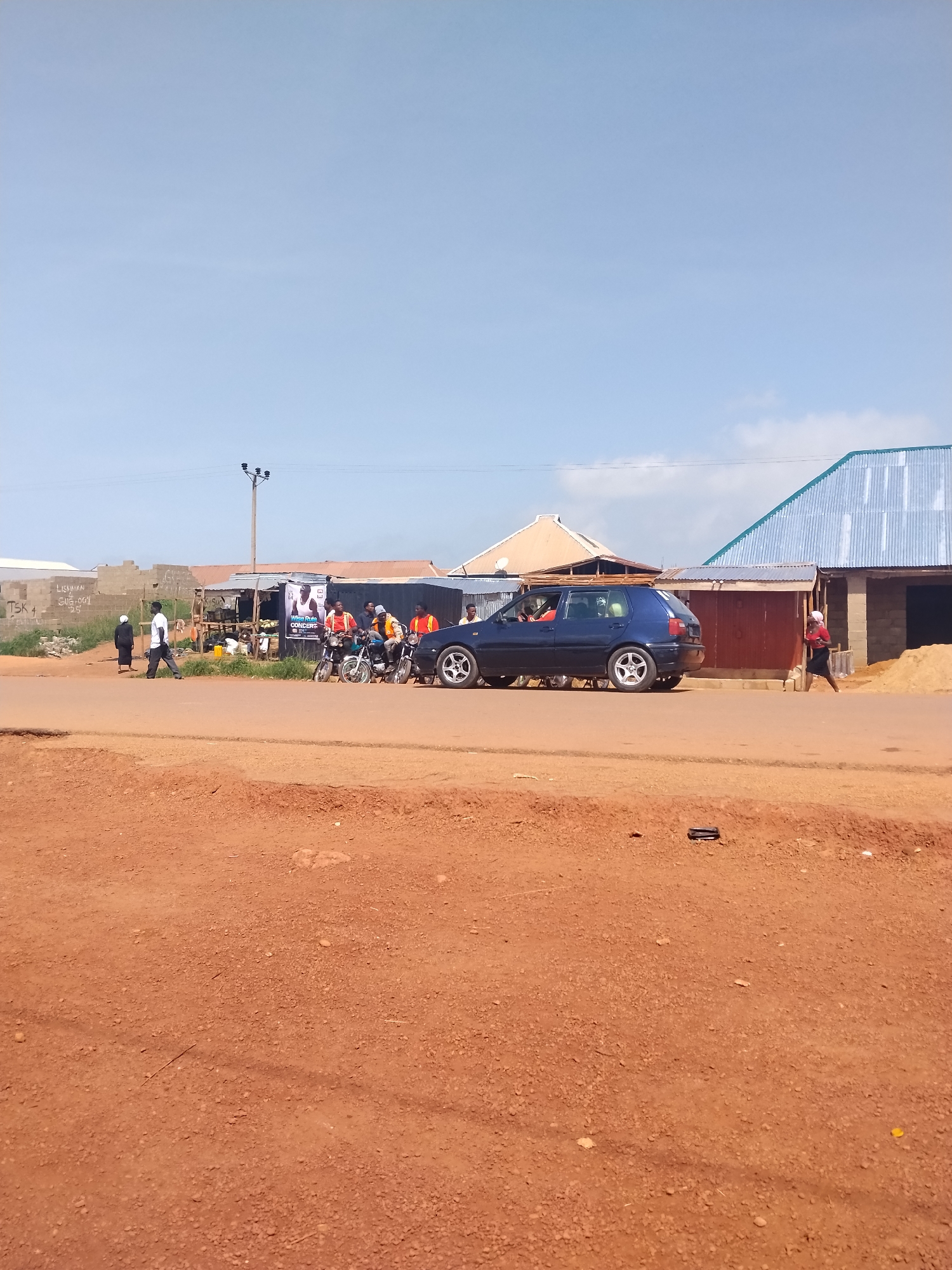 The image depicts a street scene in Durumi, Nigeria, under a clear blue sky. In the foreground, a dirt road with reddish-brown soil dominates the lower half of the frame. A dark blue hatchback car is driving on an asphalt road in the middle ground, with several people on motorcycles to its right. To the left of the car, a man in a white shirt and dark pants walks away from the camera. Behind the car and motorcycles, there are several market stalls and buildings, some with corrugated metal roofs. A poster advertising a concert is visible on one of the stalls. A woman in a red shirt and black skirt is walking across the road to the right of the car. In the background, a power pole with wires is visible, and further back, more buildings and a dusty landscape extend to the horizon. The overall impression is one of a bustling roadside marketplace in a Nigerian town.