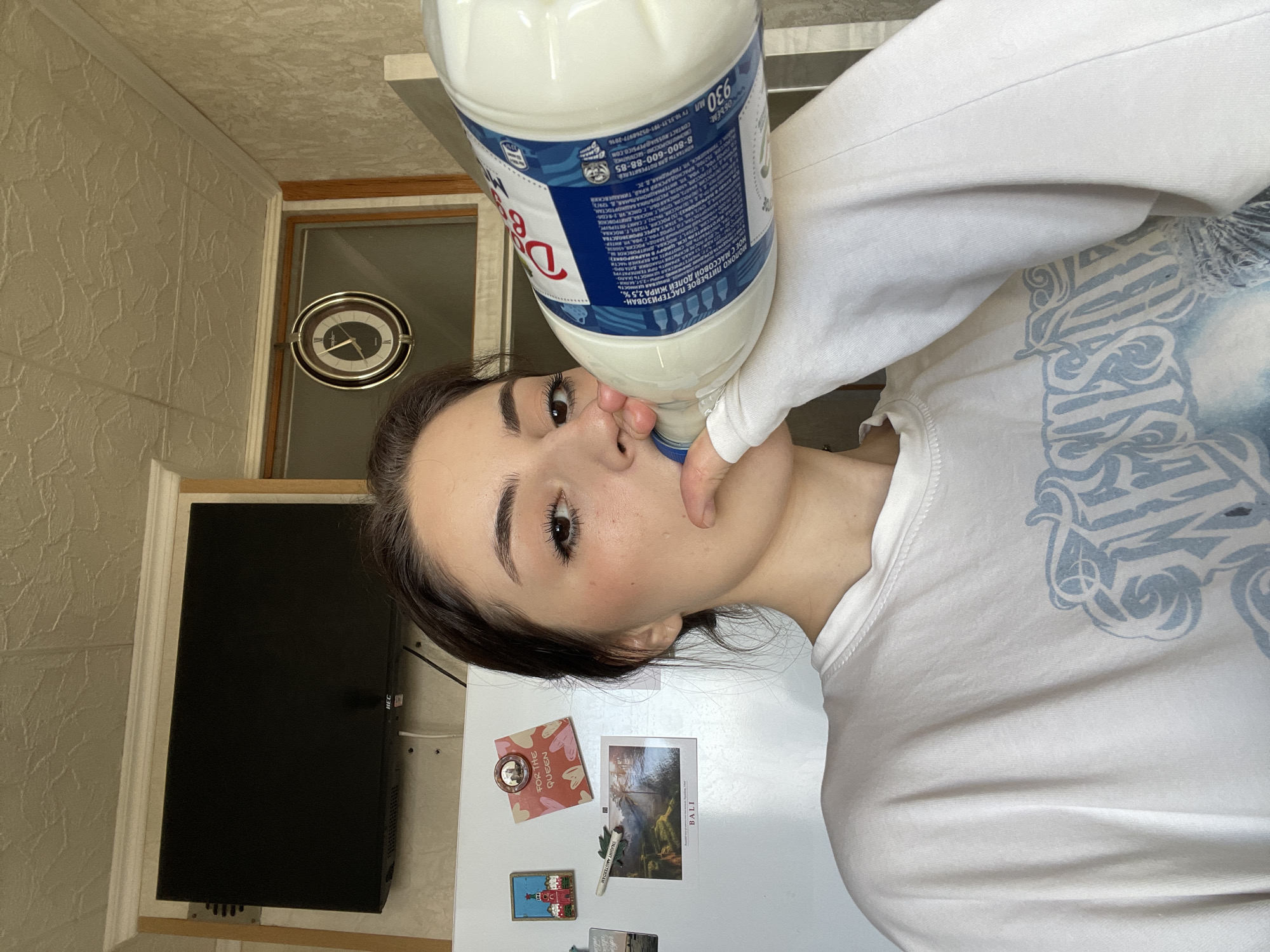 A young woman drinks directly from a large carton of "Dairy 69" milk in what appears to be a kitchen. The carton is held at an angle, tilted towards her mouth. A TV is mounted on the wall. Postcards or refrigerator magnets are affixed to the refrigerator. These include a "For the Queen" magnet, a "Bali" postcard, and a magnet depicting a "Joint Amsterdam" scene. The environment also includes a clock mounted on the wall. The carton's label indicates that the manufacturing plant is located in Russia, at the address: ROSSIYA 2, CORTOSTAN, 201, G. MOSKVA, OSAVA DMIT, ITROVSKOE. Additionally, it indicates that the product is milk with 2.5% fat content and an energy value of 120 kcal.