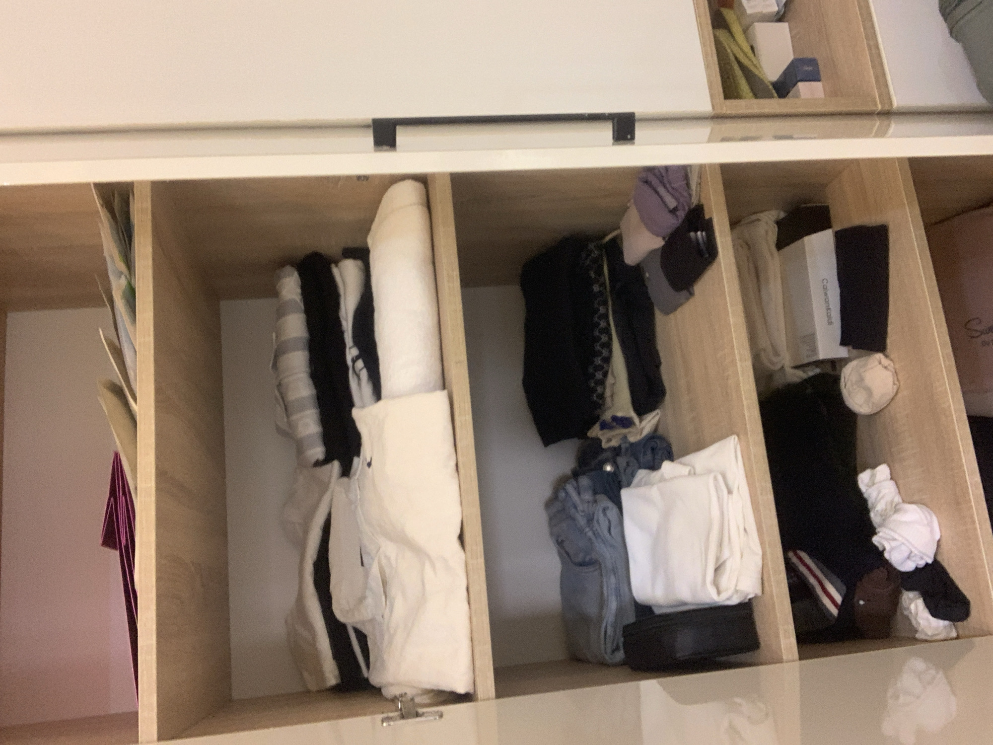 The image shows the interior of a wardrobe with multiple shelves. The shelves are filled with folded clothes, including shirts, pants, and other garments. On the top shelf, there are some folded items that appear to be papers or folders. The middle shelves contain neatly folded stacks of clothing in various colors, primarily white, black, and denim blue. The bottom shelf also has folded clothes, along with some loose items like socks and a round object that might be a ball of yarn or a cosmetic product. The wardrobe appears to be made of light-colored wood. The lighting suggests an indoor setting. The image does not provide any information about the location of Abuja, Nigeria.