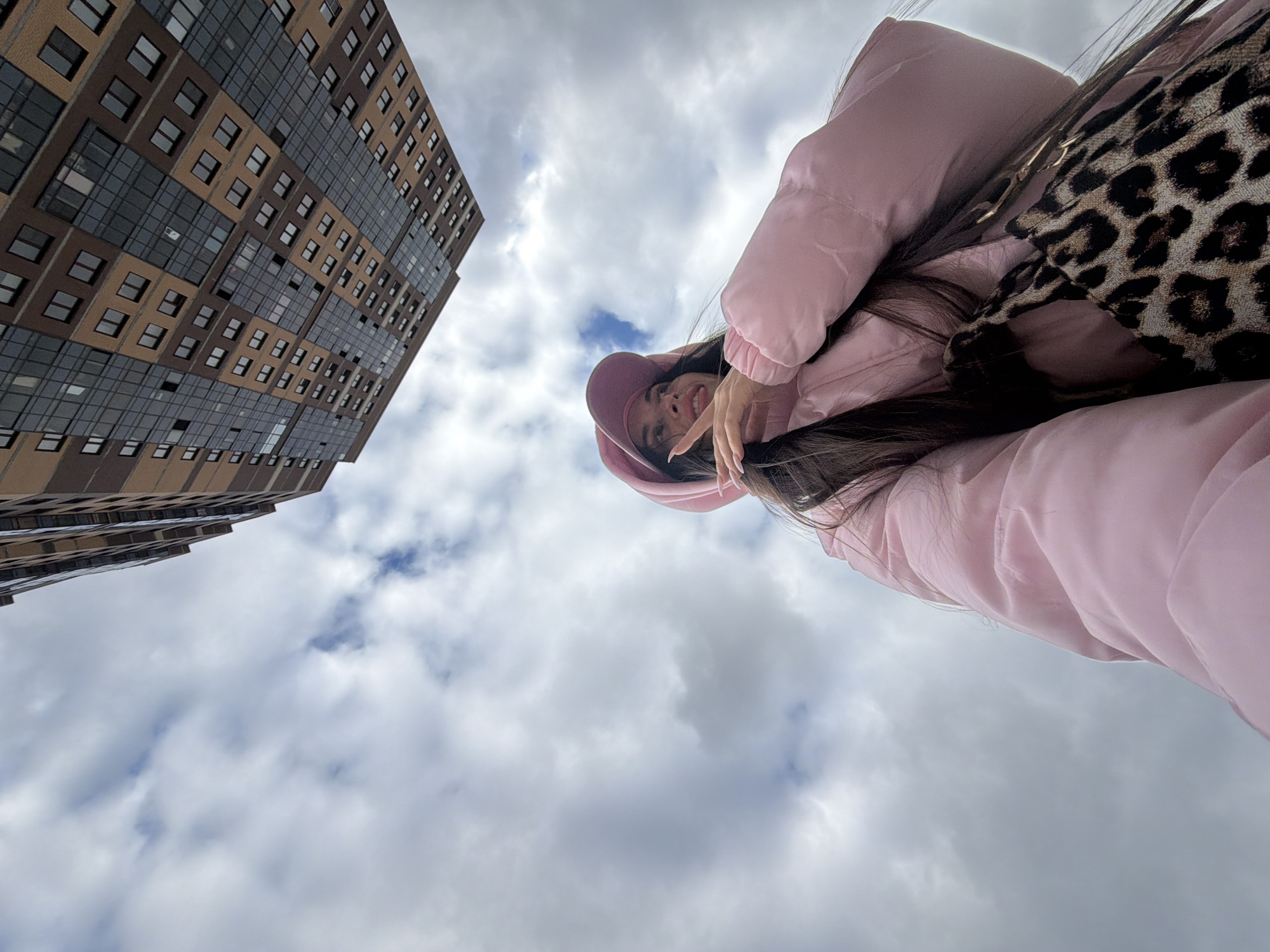 A low-angle photograph captures a smiling young person against the backdrop of a modern building and a cloudy sky in Saint Petersburg, Russia. The individual, likely female, has long dark hair and is wearing a light pink padded jacket and a matching pink hat or hood. She is positioned in the upper right quadrant of the frame, looking down towards the camera with a cheerful expression, her hand raised near her chin. A leopard-print accessory, possibly a bag or scarf, rests on her shoulder.
To the left, a tall, contemporary building rises upwards, showcasing a distinctive facade. It features a patterned mix of brown and beige rectangular panels alternating with large sections of dark, reflective glass windows, creating a gridded appearance. The architecture suggests a modern urban setting.
The sky, filling the majority of the frame, is overcast with a mixture of grey and white clouds, indicating daytime. Small patches of blue sky are visible peeking through the clouds, particularly towards the center. The perspective is from below, looking up, making the building appear imposing and the person slightly elevated. No visible text is present in the image.