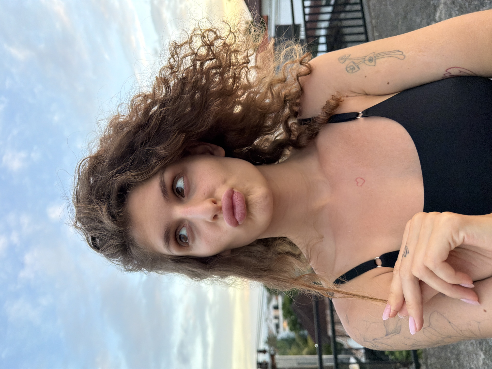 A young woman with light brown curly hair is posing for a selfie with a "duck face" expression. She is wearing a black top. Her arms are visible, with a few tattoos on her upper arms. The background shows a cloudy sky, a few buildings, and a metal railing. The location is North Kuta, Indonesia.