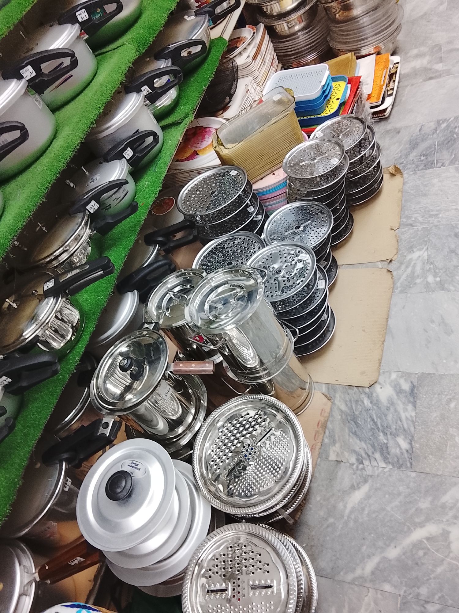 The image depicts a store display in Gulgasht Colony, Pakistan, featuring a variety of kitchenware. Stainless steel pots and pans with black handles are arranged on tiered shelves covered with artificial green turf. Adjacent to the cookware are stacks of metal graters and strainers. Further back, there are organized stacks of colorful plastic containers and plates. The background shows additional kitchen supplies. The floor is marble.