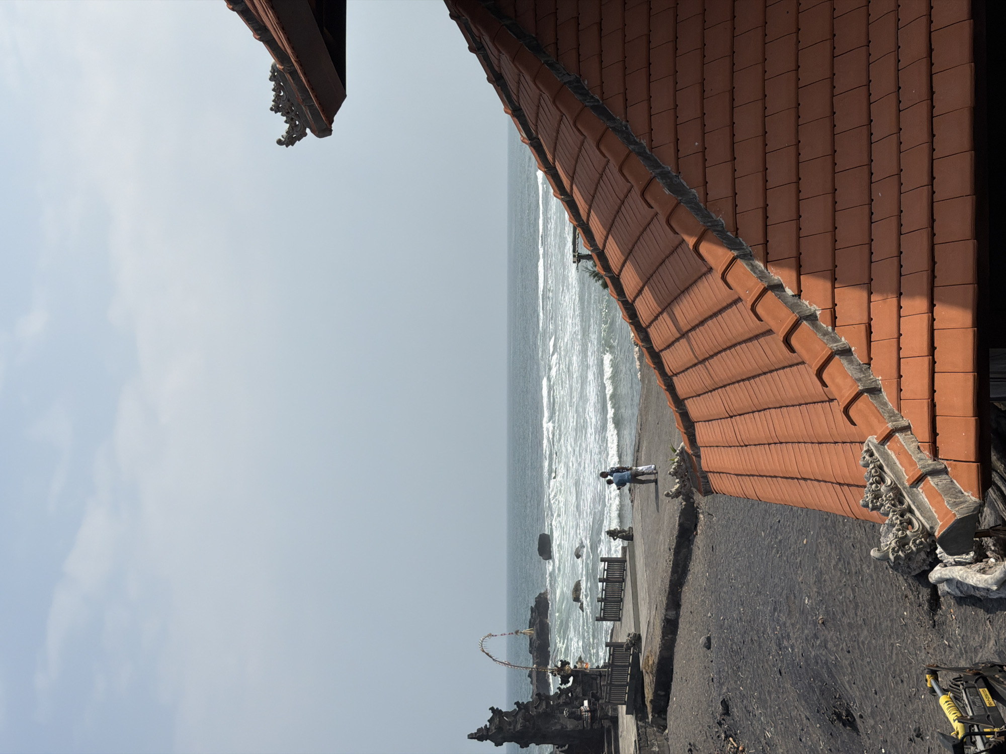 The image shows a coastal scene in Mengwi, Indonesia, with a traditional Indonesian tiled roof in the foreground. The ocean is visible in the background, with waves breaking on a dark sand beach. There are a few people standing on a paved area near the water, and a stone structure with intricate carvings is visible on the left side of the frame. The sky is overcast.