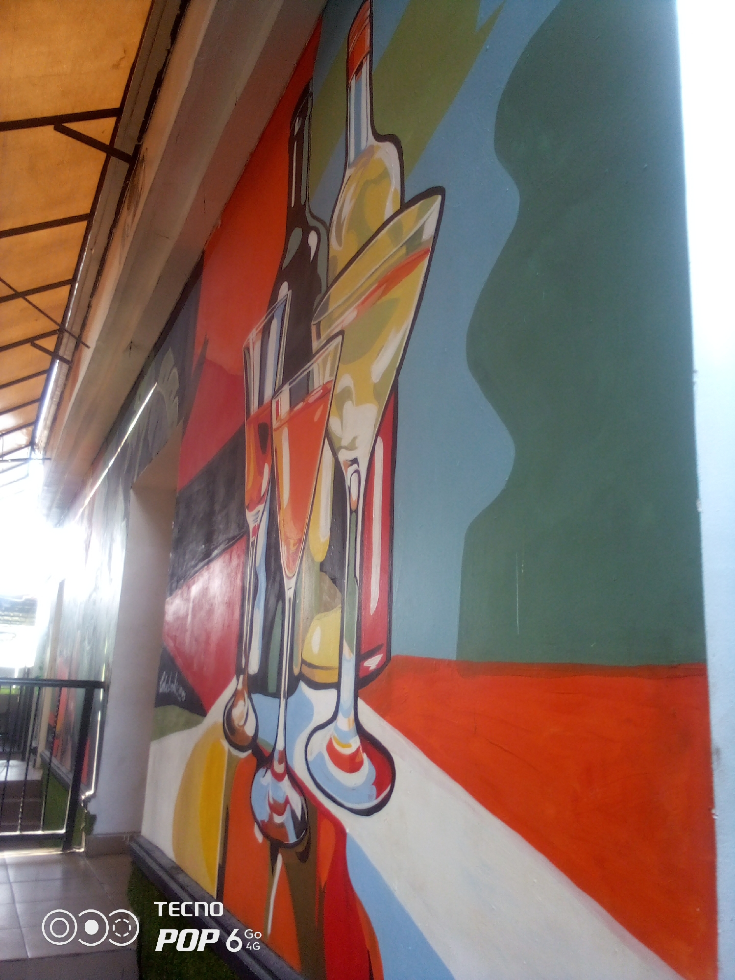 This is a photograph of a vibrant mural on an exterior wall in the city of Howayi, Nigeria. The mural features an abstract, stylized depiction of bottles and glasses, likely representing drinks. The artwork employs bold colors, including shades of red, orange, yellow, blue, green, and black, with distinct outlines. In the foreground, the mural shows multiple glasses, some filled with a reddish-orange liquid, and what appear to be bottles. In the background, there are broader strokes of color and shapes. To the left of the mural, a metal railing and stairs are visible, suggesting this is an outdoor area, possibly a walkway or entrance. Above the mural, a portion of a yellow awning supported by dark metal beams is visible. The overall impression is one of a lively, artistic decoration on a building in a public space. The bottom right corner of the image displays a "TECNO POP 6 4G" watermark.