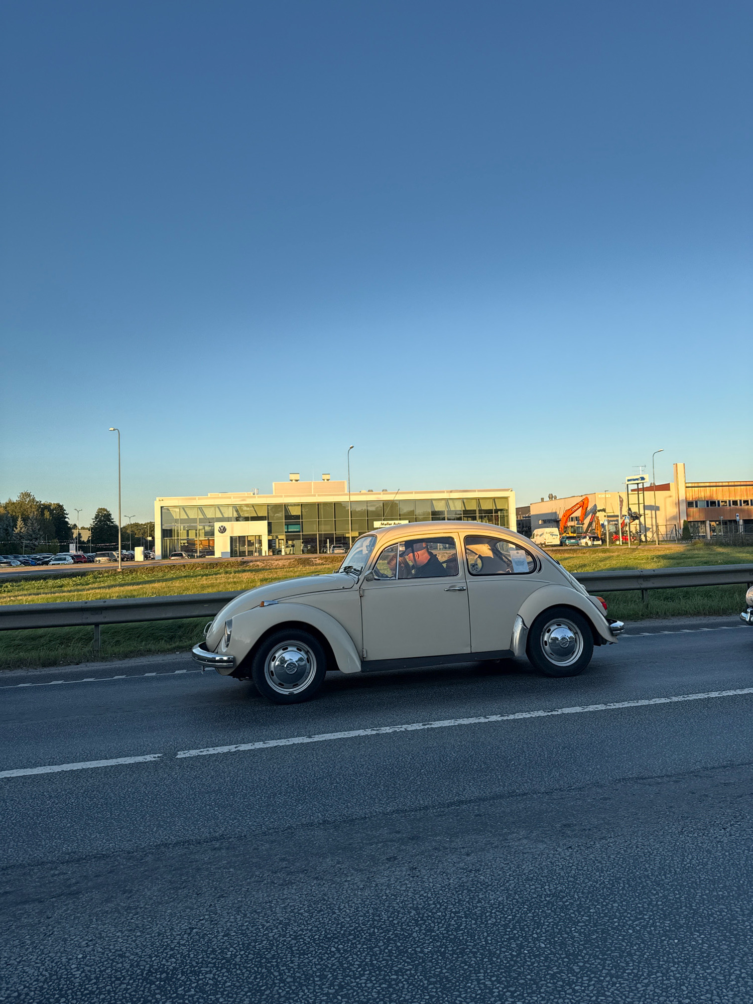 A beige Volkswagen Beetle drives on a road in Peetri alevik, Estonia. The car is in motion, facing right. In the background, a modern building with large glass windows stands behind a metal fence and grassy median. A bright blue sky with no clouds is visible above. Construction equipment, including an orange excavator, is present near the building. Streetlights and trees are also visible on the left side of the image. The road is paved and has white lane markings.