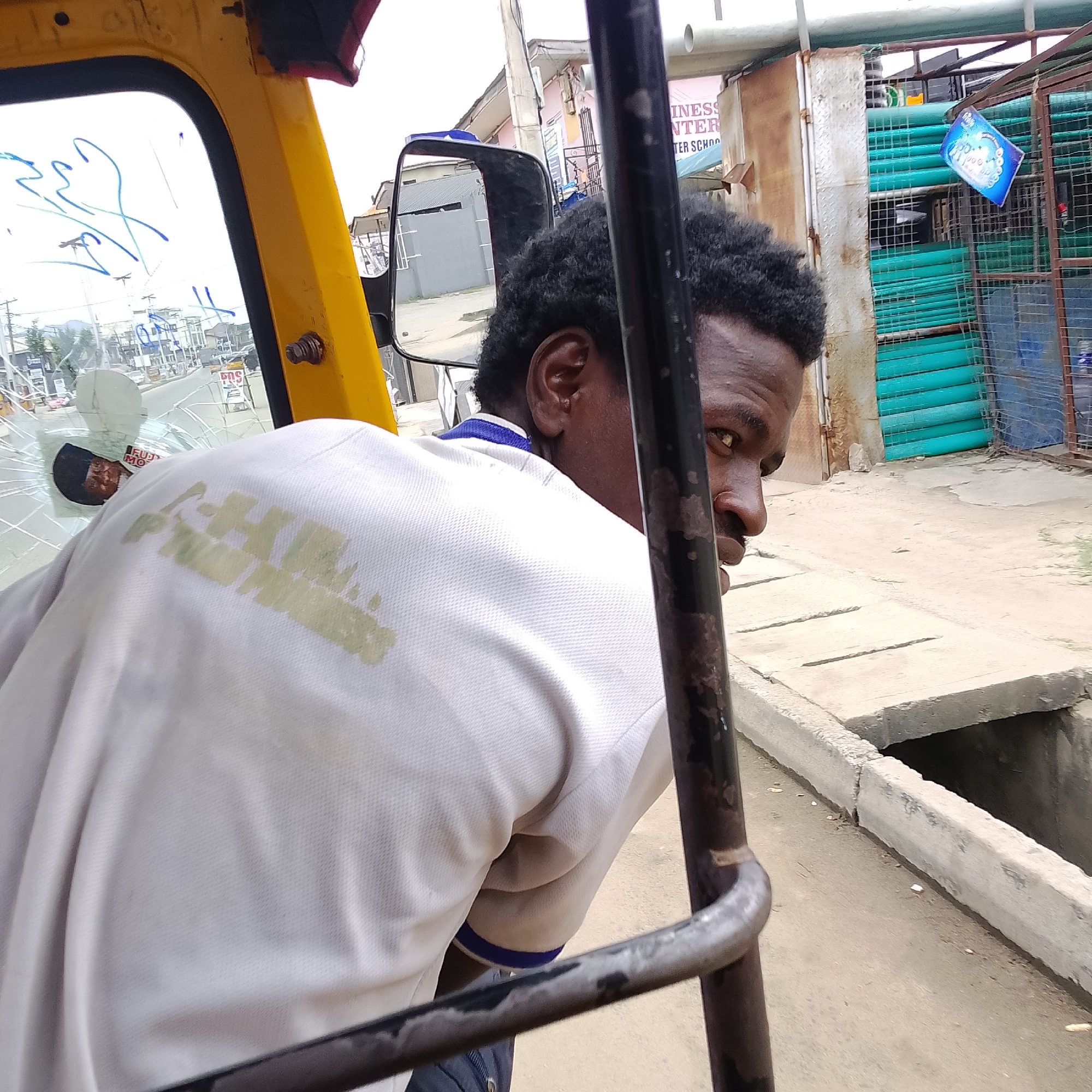 This is a medium shot, eye-level, outdoor, horizontal, eye-level photograph taken from inside a yellow auto-rickshaw in Ado, Nigeria. The focus is on the upper body of a Black man looking over his right shoulder towards the camera. He has short, dark, curly hair and is wearing a light-colored polo shirt with a blue collar. His left ear and part of his face are visible, with his right eye looking directly at the camera.
The foreground is dominated by the interior of the auto-rickshaw, including a black metal pole in the center of the frame and the yellow body of the vehicle on the left. The window of the auto-rickshaw is visible on the left, with some blue scribbles on it.
The background shows a street scene. To the right of the man, a side mirror of the auto-rickshaw reflects a street with buildings and some vehicles. Beyond that, a fenced area contains stacked green pipes. A blue and white sign is attached to the fence. Further in the background, buildings and utility poles are visible. The ground appears to be paved concrete. A concrete drain or culvert is visible in the lower right portion of the frame. The lighting suggests it is daytime, with overcast or diffused light.
