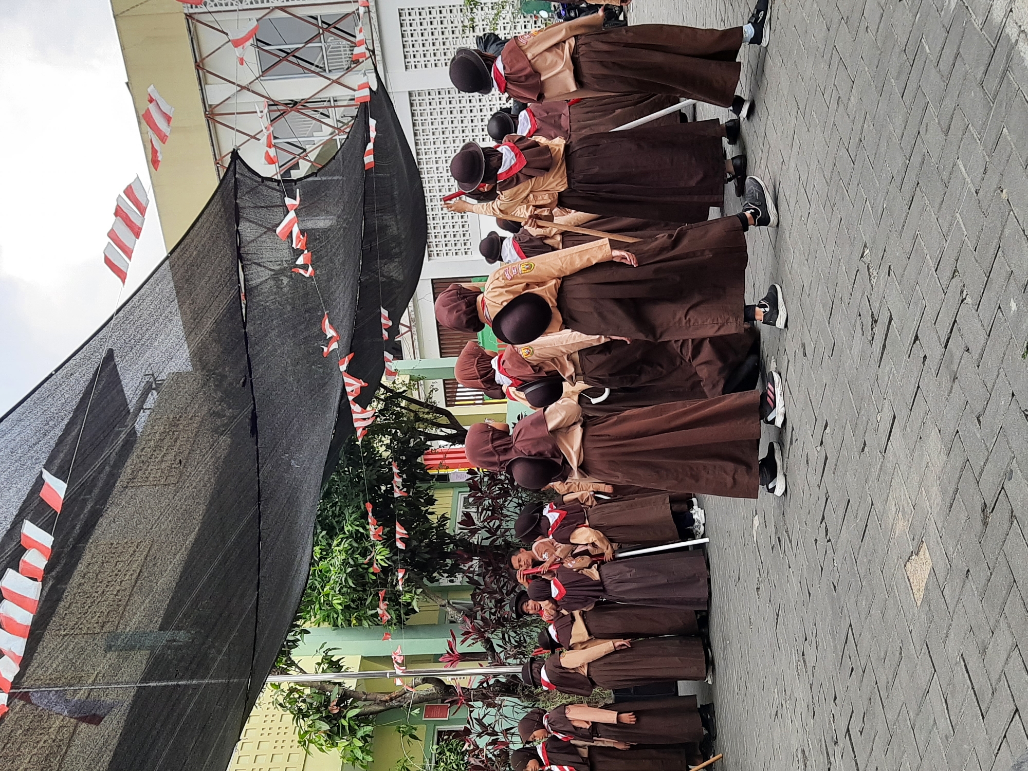 In Cimenyan, Indonesia, a group of young people, appearing to be students, are participating in an outdoor event. They are dressed in matching uniforms consisting of brown long skirts and tan long-sleeved shirts, with some wearing brown headscarves and hats. Many are holding long white poles. Red and white Indonesian flags are strung overhead, decorating the area. The event is taking place on a paved courtyard, with buildings and trees visible in the background. The students are in a formation, suggesting they are engaged in a parade or ceremony.