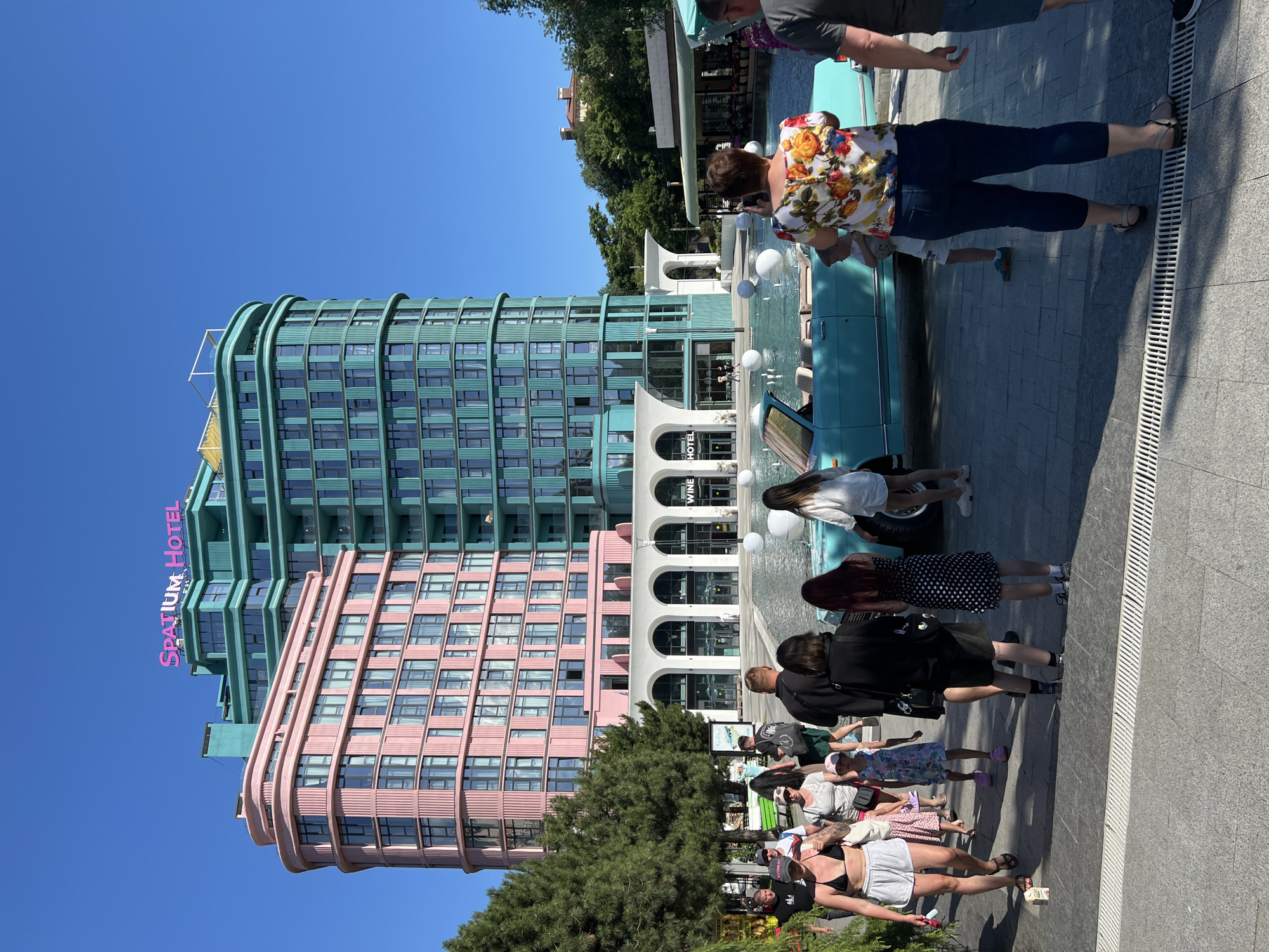 The photograph captures the "SPATIUM Hotel" building in Odesa, Ukraine, characterized by its distinctive pink and teal colored architecture. In the foreground, a group of people are walking on a paved area. A light blue convertible is parked near a water feature with floating white spheres. The hotel building, with its curved design, stands in the background under a clear blue sky. The scene suggests a public area, possibly a promenade or plaza, adjacent to the hotel.
