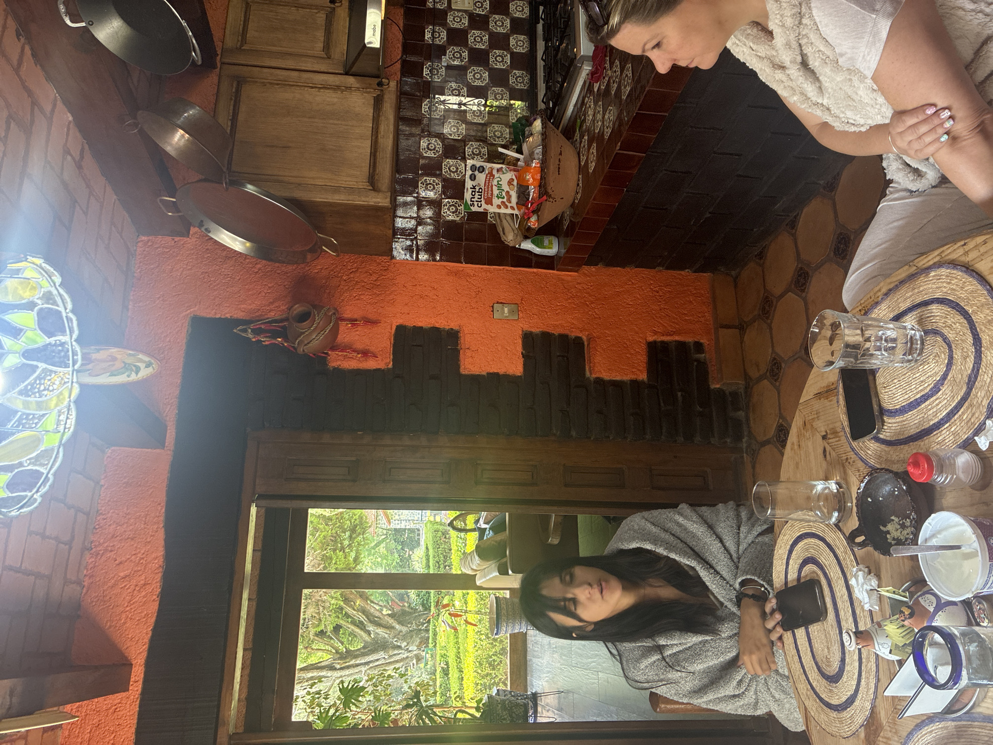 Two women are at a round table in a room with a window overlooking a garden. One woman wears a light-colored shirt and light-colored fleece vest. The other woman wears a grey robe. The table has straw mats and several glasses. One cell phone lays on the table. Pots and pans hang on the wall. The wall is decorated with orange, black and brown tiles. The garden has plants and trees and the outdoors seems to be in the city of Cañada de Madero, Mexico.