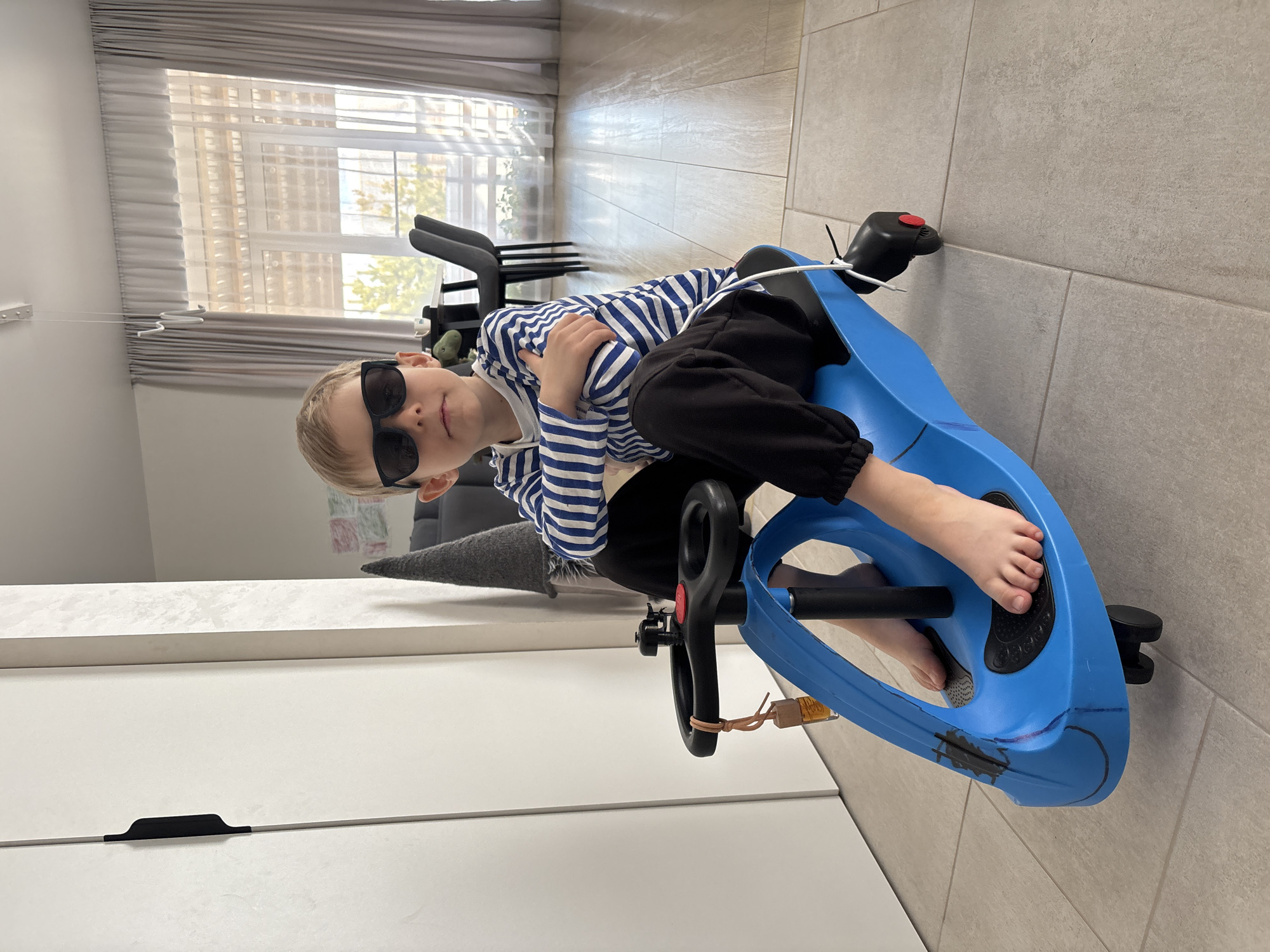 A young boy is depicted indoors, sitting confidently on a blue ride-on toy, likely a drift trike. He is wearing dark sunglasses, a blue and white horizontally striped long-sleeved shirt, and black pants. His arms are crossed over his chest, and he looks directly at the camera with a calm and composed expression. His bare feet are visible, resting on the black footrests of the toy.
The blue ride-on toy features black handlebars with a red button on one side, and black footrests. It shows signs of use, including some black markings and a red line drawn on its surface, and a light-colored rubber band or string tied near the steering mechanism.
The setting appears to be a modern home or apartment in Chaikovsky, Russia. The room has a light-colored tiled floor and white walls. In the background, large windows with sheer white and darker grey pleated blinds allow natural daylight to flood the space. Parts of dark-colored chairs and a grey cushion are visible behind the boy. To the left, a white cabinet or dresser with a black horizontal handle can be seen. The environment suggests a clean and well-lit interior. No visible text is present in the image.