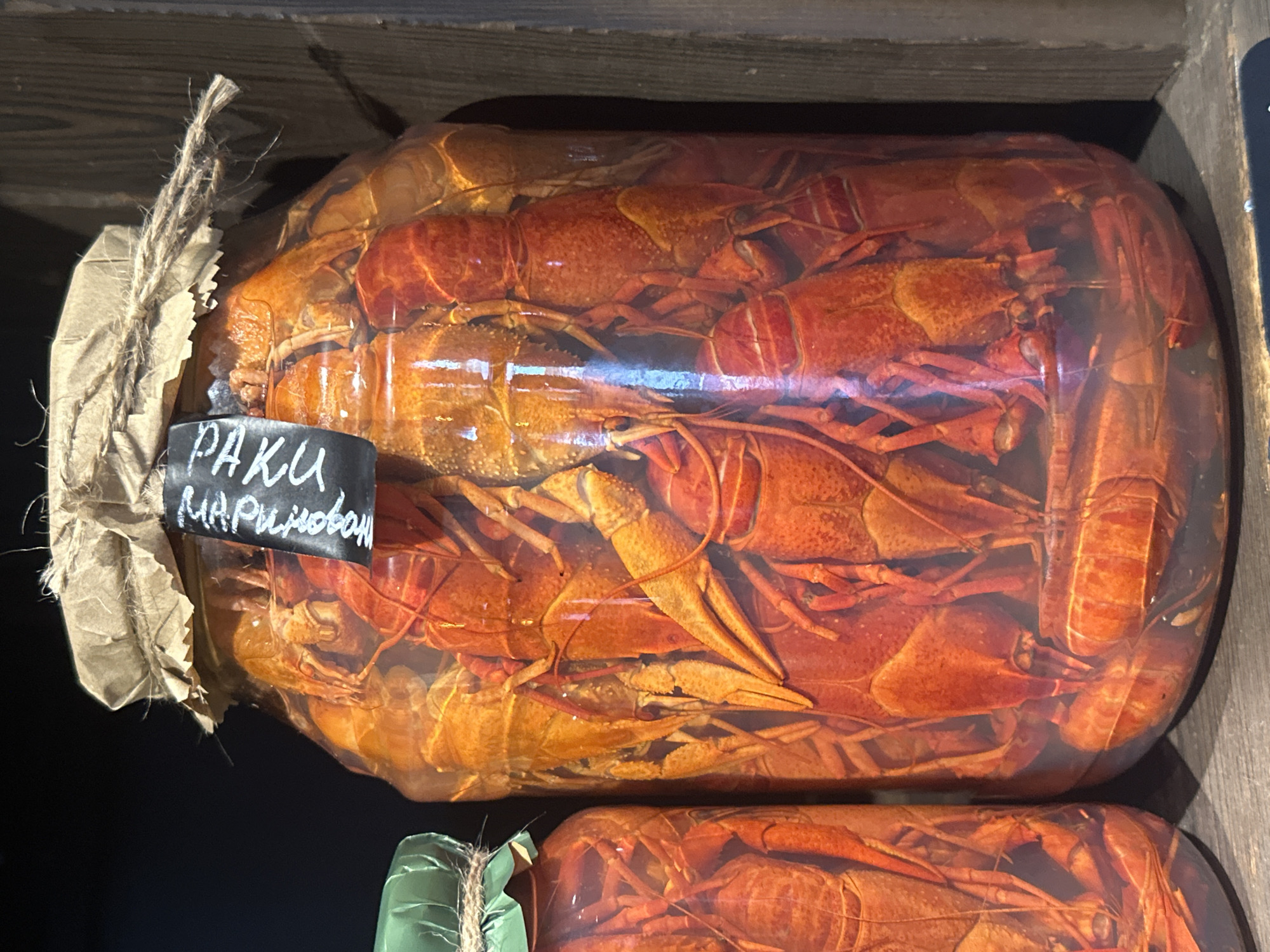A large glass jar filled with cooked crayfish sits on a wooden shelf. The jar is labeled with the word "PAKU" in Cyrillic letters, followed by "MAPИНOBOH." The top of the jar is covered with brown paper and tied with twine. Another similar jar, also containing crayfish, is visible beneath the first one. The location is Новоглаголево, Russia.