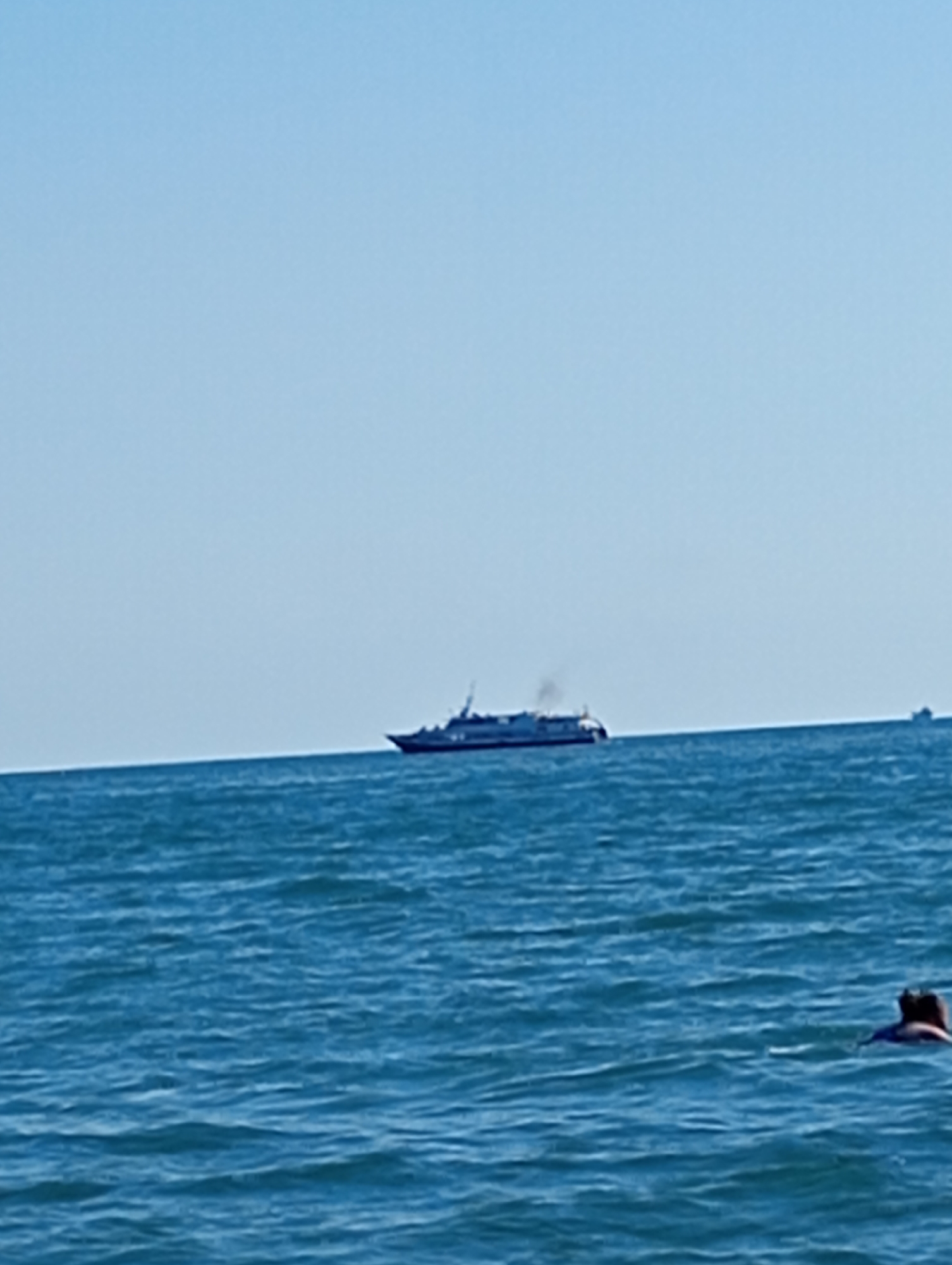 The image captures a serene maritime scene under a clear blue sky, likely depicting the waters off the coast of Sokhumi, Abkhazia. In the distance, a sleek white passenger hydrofoil with dark blue accents cuts through the calm blue sea, leaving a faint trail of smoke from its engines. Closer to the viewer and slightly to the right, a person is partially visible in the water, appearing to be swimming or wading, with their head and shoulders above the surface. The vast expanse of the ocean dominates the foreground and midground, with gentle ripples indicating calm conditions. The horizon line is sharp, separating the tranquil water from the cloudless sky. Another, smaller vessel is faintly visible on the horizon to the right of the hydrofoil.