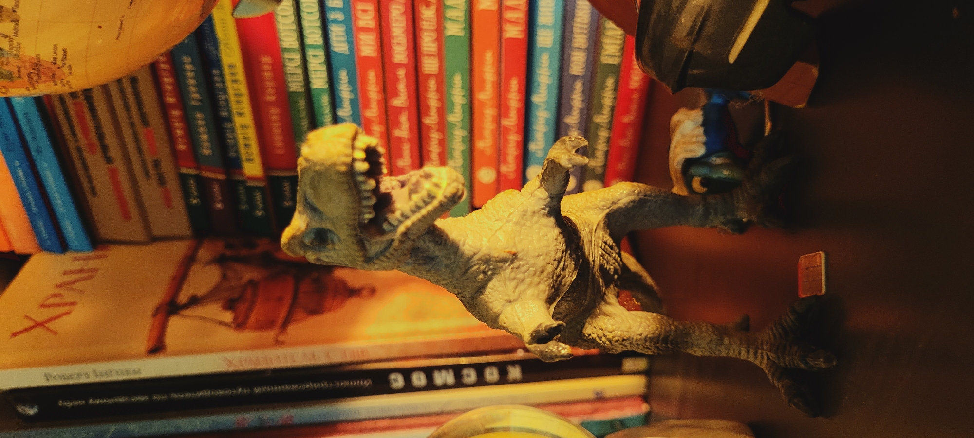 A toy Tyrannosaurus Rex stands on a dark surface in front of a stack of books and a globe. The dinosaur is light green and has its mouth open, showing its teeth. The books are of various colors, and some have titles in a foreign language. The globe is partially visible on the right side of the image. The lighting is dim, casting shadows and highlighting the dinosaur. The scene appears to be set indoors, possibly on a bookshelf or desk.