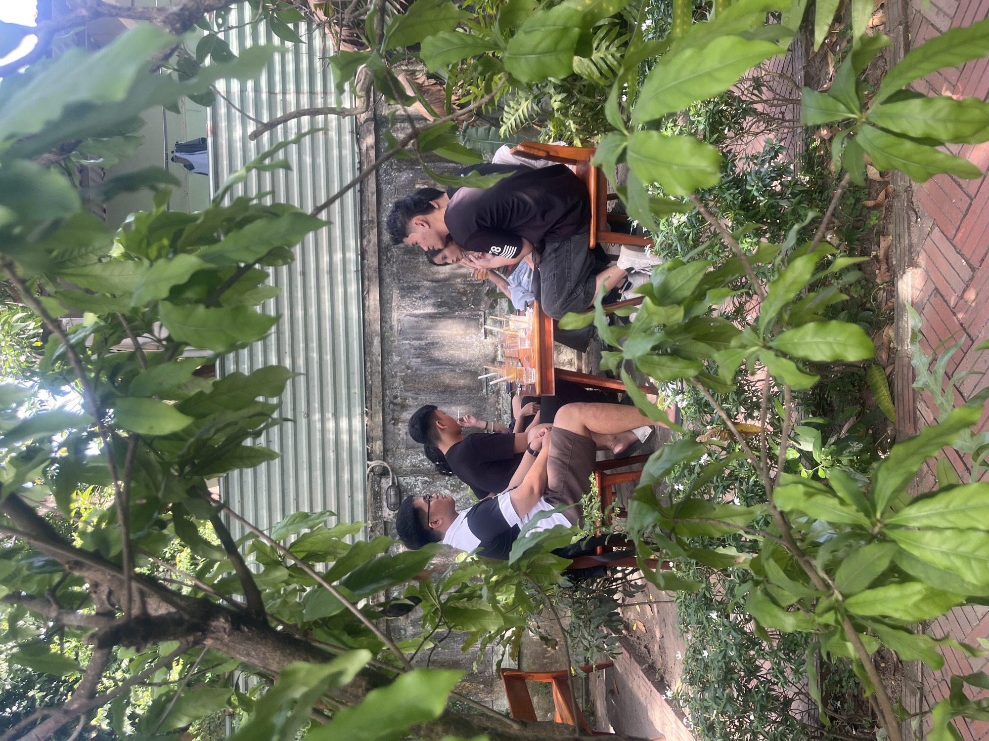 The image captures an outdoor social gathering of four young adults at a cafe or garden restaurant, likely situated in Đà Nẵng, Vietnam. The scene is viewed through a dense natural frame of green leaves and tree branches in the immediate foreground and midground.
Four individuals are seated around a square, light-colored wooden table. Multiple glasses filled with beverages and straws are present on the table.
From the left, a male in a black and white short-sleeved polo shirt and dark shorts is seated on a wooden chair, facing right towards the group. He has short dark hair and glasses.
Adjacent to him, another male, wearing a black t-shirt, holds a cigarette in his right hand. He is also seated, facing right.
Across the table, a female in a light-colored top is partially visible, facing left, engaged in conversation.
On the far right, a fourth male, wearing a black t-shirt with a white '78' graphic on the sleeve and dark trousers, is seated facing left, looking towards the center of the group. He wears white sneakers.
The ground consists of red brick paving laid in a herringbone pattern, interspersed with green plants and small white flowers. Lush green foliage, including shrubs and trees, surrounds the seating area. In the background, behind the group, a textured, light grey concrete wall shows signs of weathering. Above this wall, a light grey corrugated metal fence or structure is visible, with articles of clothing, including a white shirt, hanging on a line above it. The scene is illuminated by natural daylight.