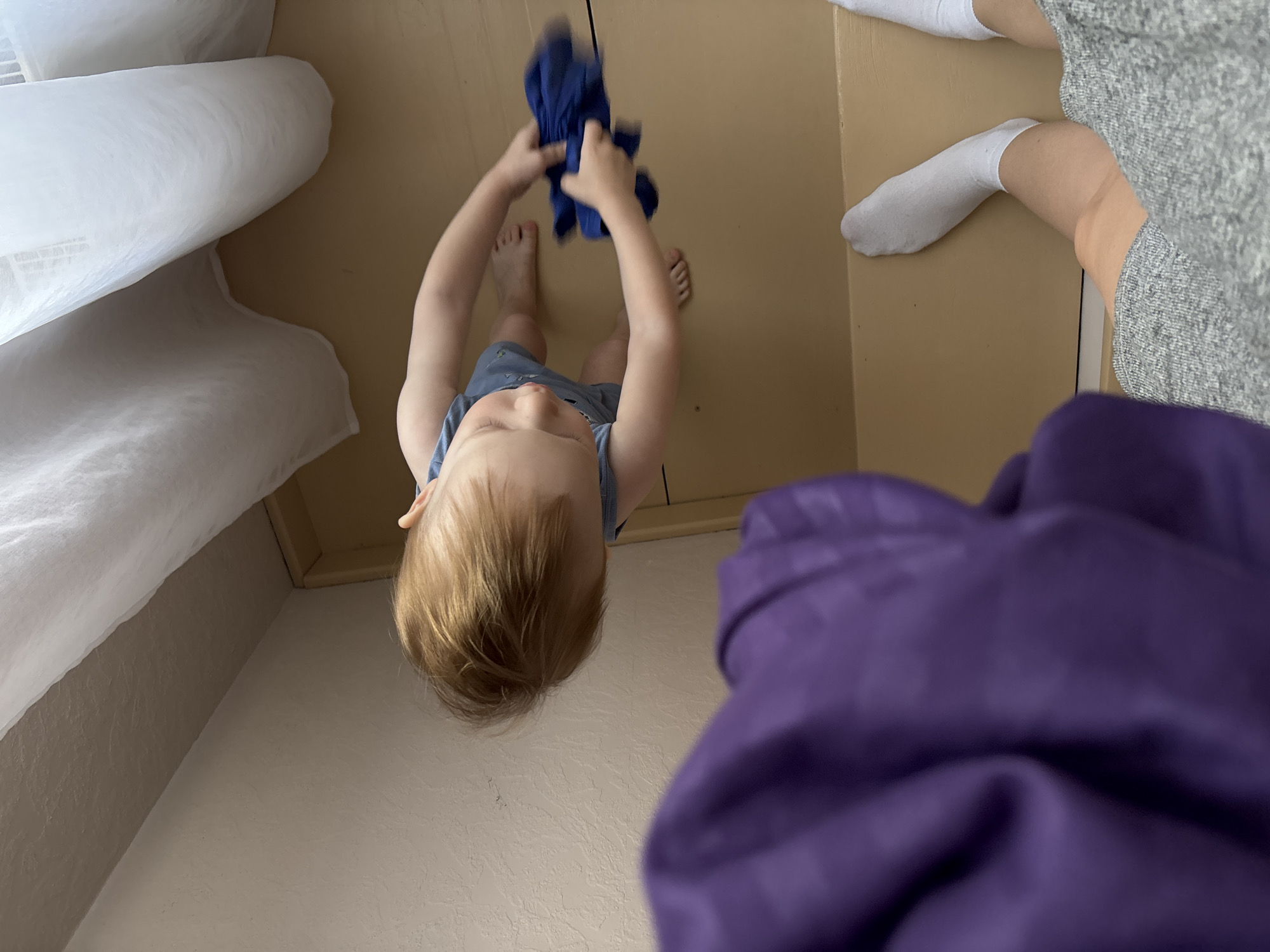 A high-angle shot shows a child standing barefoot on a light brown surface, holding a blue object above their head. The child wears a blue garment. A white object is in the upper left corner. To the right of the child, an adult figure is partially visible, wearing white socks and a gray garment. A purple object is in the foreground, partially obscuring the view. The scene appears to be indoors. The location is Ферма, Russia.