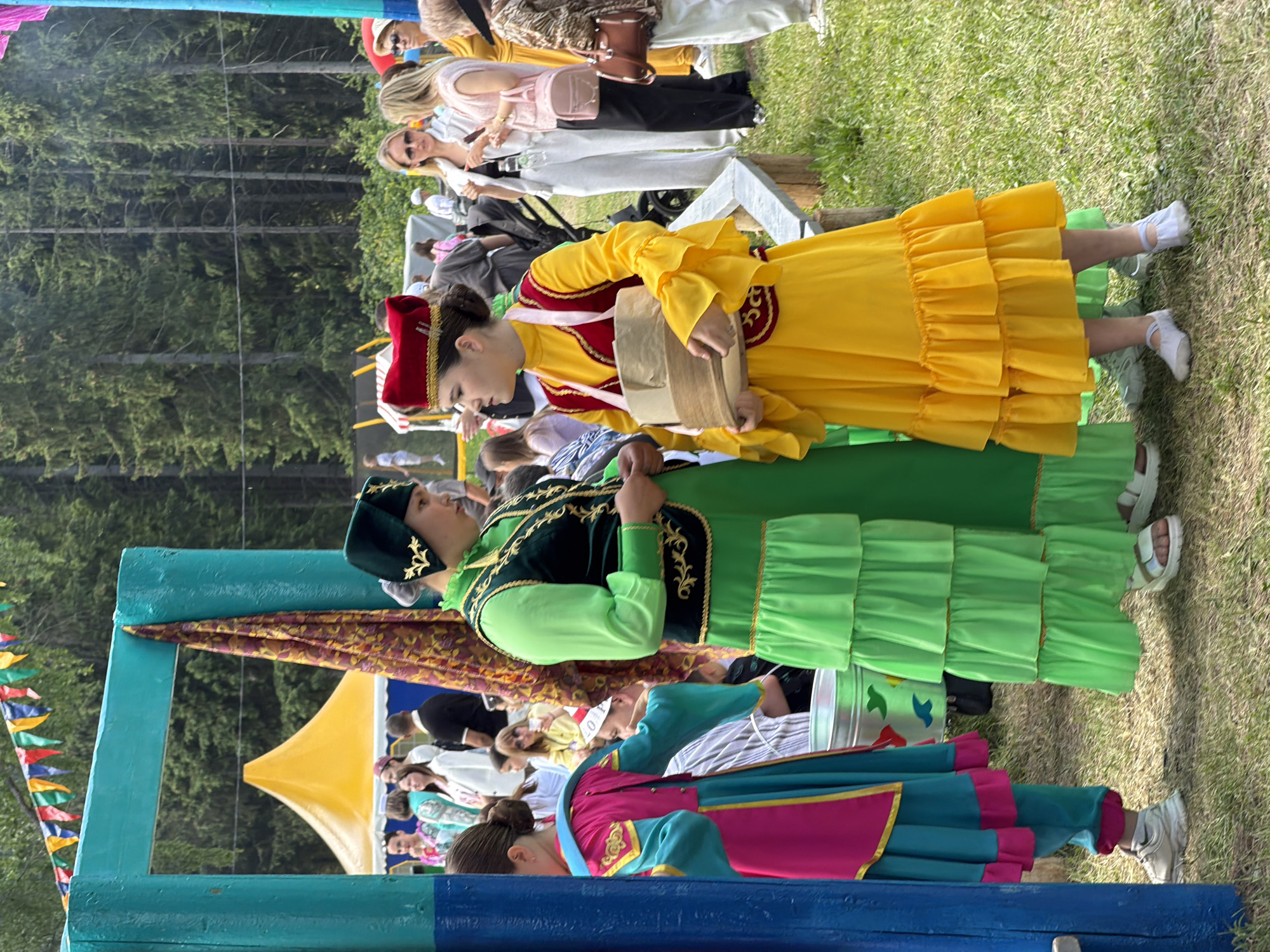 The image captures an outdoor daytime scene at a cultural gathering. In the foreground, two young women stand on green grass. The woman on the left wears a bright green long-sleeved top, a tiered green skirt, a dark green sleeveless vest with gold embroidery, and a dark green hat with gold embellishments; she wears white platform sandals. The woman on the right wears a bright yellow long-sleeved dress with ruffled sleeves and a tiered yellow skirt, a red sleeveless vest with gold embroidery, and a red hat with gold embellishments. She holds several light brown paper bags against her torso and wears white ballet-style soft shoes. Both women appear to be interacting.
To their left, a prominent turquoise-blue wooden archway stands, with a patterned maroon, yellow, and green fabric draped over its horizontal beam. The lower section of the left archway post is painted darker blue. To the far left, partially visible, is a young person in a pink and blue traditional-style outfit with white sneakers, facing away. A metal bucket with colorful fish decals is on the ground near this person. In the midground, behind the main subjects, several adults are visible, including a woman in a white pantsuit and sunglasses, and another in a light-colored top with a pink handbag. A yellow conical tent is partially visible behind the archway on the left. The background features a dense forest of tall evergreen trees under natural daylight. A string of colorful triangular flags is visible across the top of the frame. The event takes place on a grassy field.