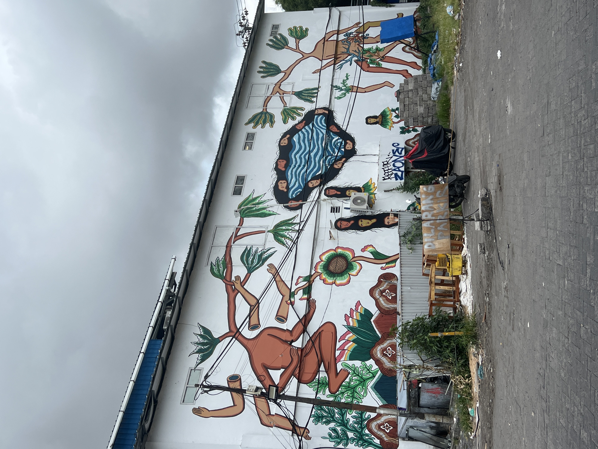A mural adorns a white, two-story building in Kuta, Indonesia. The mural features stylized, interconnected figures with brown skin tones, representing humans, flora, and water elements. A figure with tree branches emerging from its head sits amidst flowers and plants at the bottom of the mural. Above this, a pool of water with faces is depicted. Higher up are more figures with outstretched limbs. Power lines run across the mural.
In front of the mural, there is a paved street with a parked motorcycle covered with a black and red tarp, pallets, bushes, and trash. The sky is overcast.