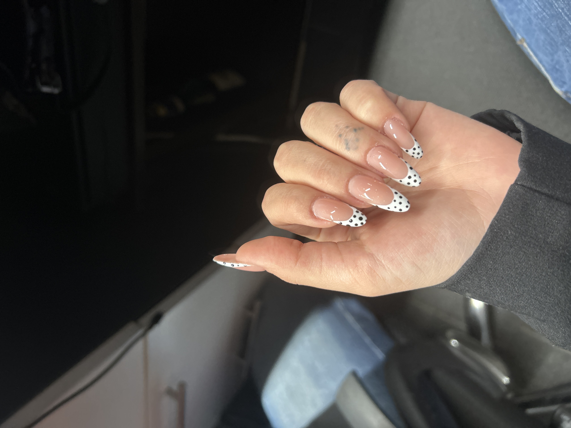 A close-up, overhead shot shows a person's hand with light skin, adorned with long, almond-shaped acrylic nails. The nails feature a French tip design with a sheer pink base and white tips, decorated with small black polka dots. A faint, dark blue tattoo is visible on the middle finger. The background is out of focus, appearing to be an indoor setting with dark and muted tones. Some objects in the background suggest furniture, possibly chairs or a desk, with hints of blue fabric, potentially denim, and reflective surfaces. The lighting is dim, creating soft shadows. No specific location in Tbilisi, Georgia, can be identified from the provided image.