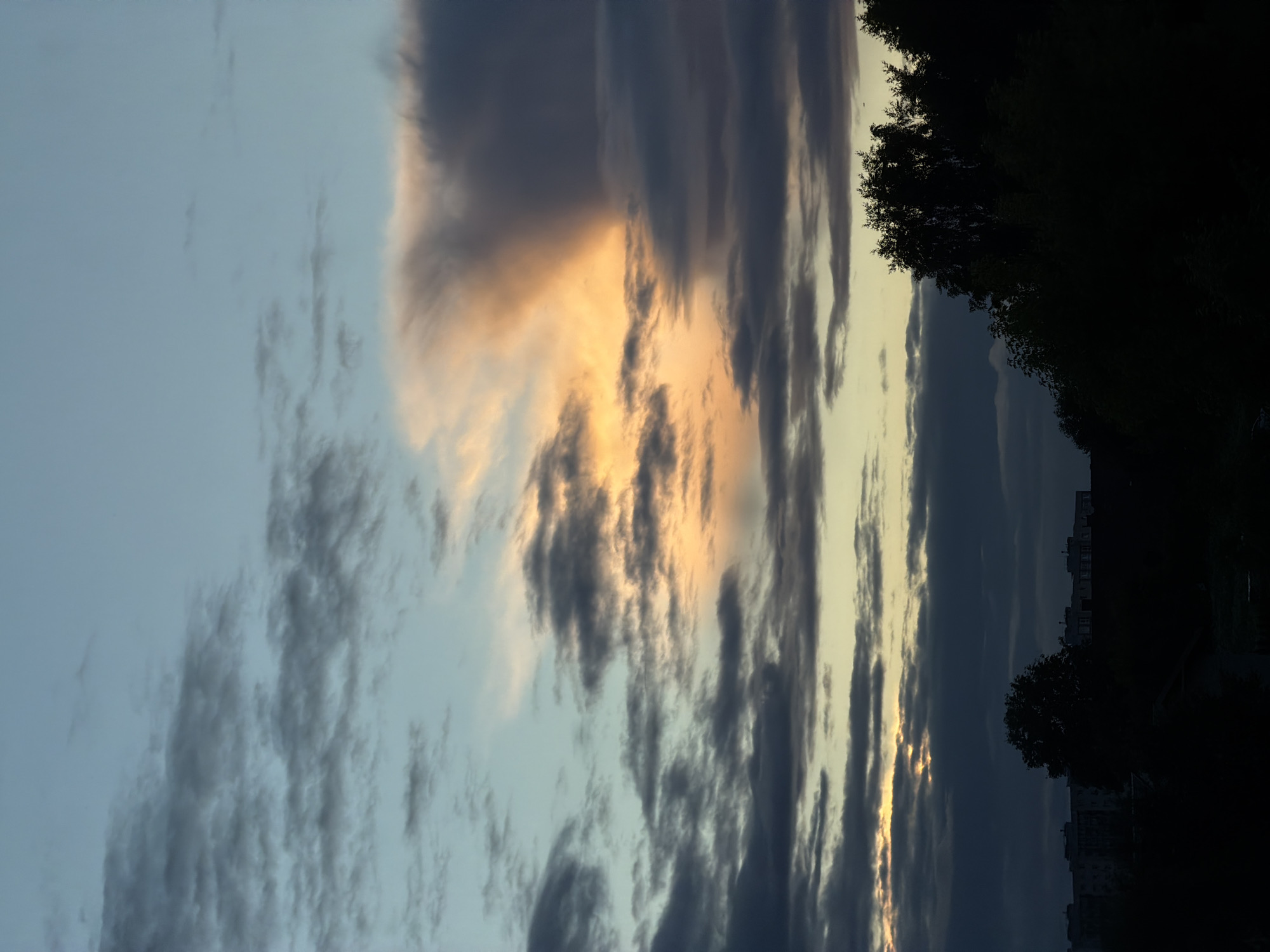 The image depicts a dusky sky above Dreiliņi, Latvia. The sky is filled with a mix of clouds, some dark and heavy, others wispy and tinged with orange and yellow from the setting sun. A large, dramatic cloud formation dominates the upper right portion of the frame, catching the light in a striking display. Below the sky, the silhouettes of trees and buildings are visible against the fading light. The overall impression is one of a serene, twilight scene in a suburban or semi-rural setting. There are no people, animals, or specific actions captured in the image. The environment is characterized by the natural elements of the sky and landscape.