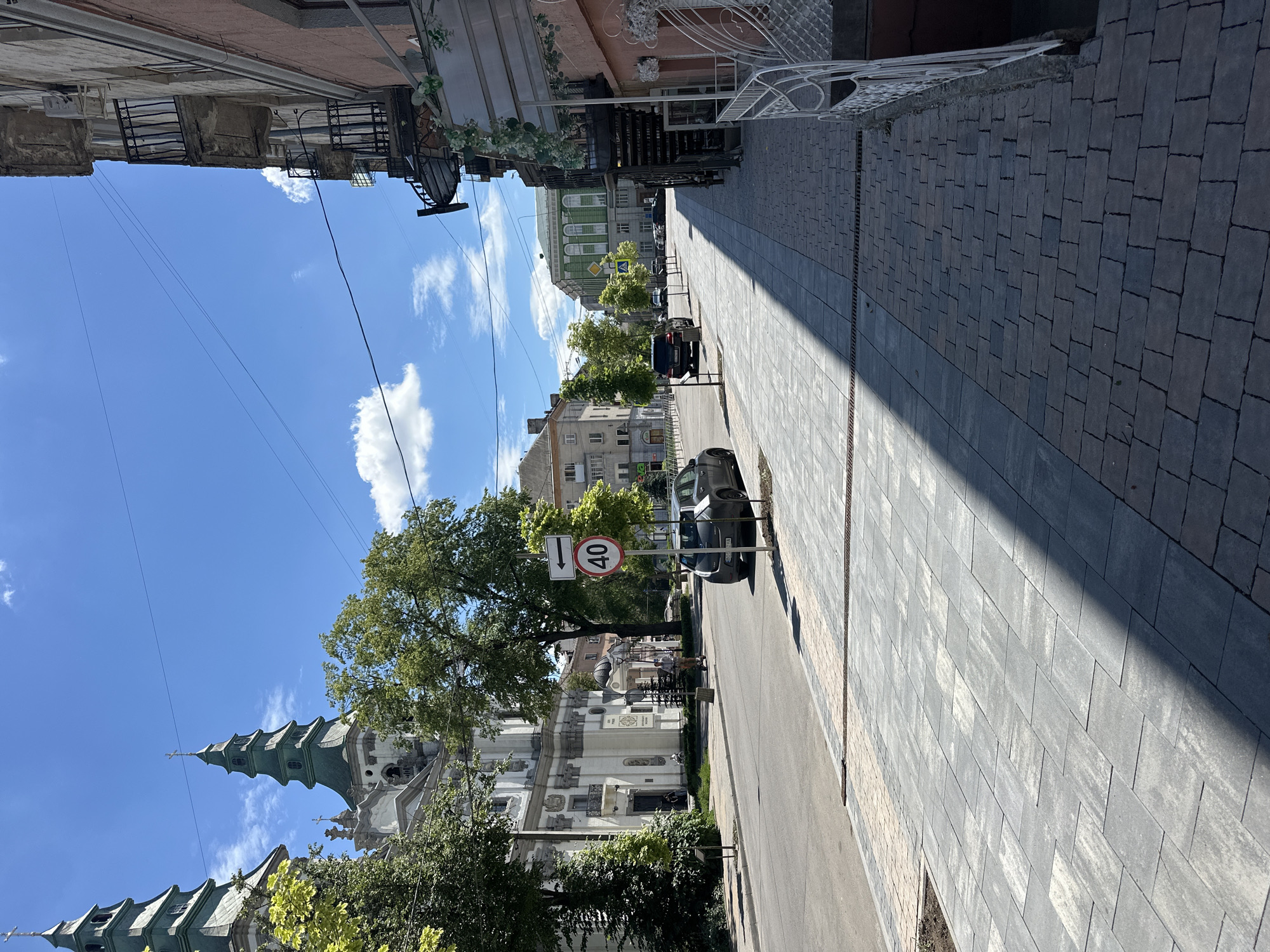 The image depicts a street scene in Ternopil, Ukraine. On the left side of the street, a large white building with prominent green spires, characteristic of a church or cathedral, stands under a bright blue sky with scattered clouds. Trees line the sidewalk in front of the building. A dark-colored car is parked on the right side of the road, and a speed limit sign showing "40" is visible near it. The sidewalk in the foreground is paved with gray and beige rectangular tiles. The right side of the image features a building with balconies and ornate railings. The street is paved with asphalt and appears to be a city thoroughfare with visible power lines overhead. The overall scene suggests a sunny day in a European city.