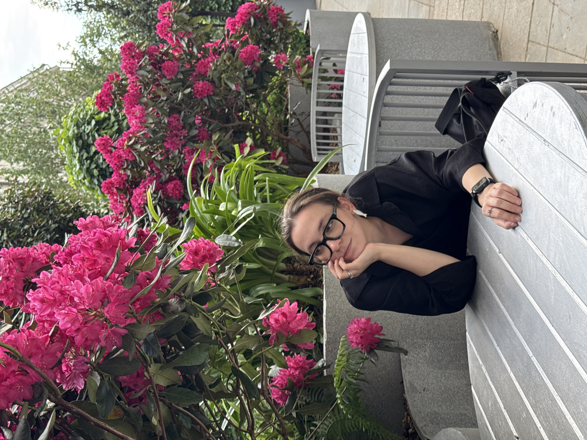 A woman with glasses and wearing a black shirt is resting her head on her hand and leaning against a grey bench. She is surrounded by vibrant pink rhododendrons and other greenery. A black bag is placed on the bench next to her. The bench has a unique design with curved, tombstone-like ends. The location appears to be outdoors, possibly a park or garden area in Moscow, Russia.