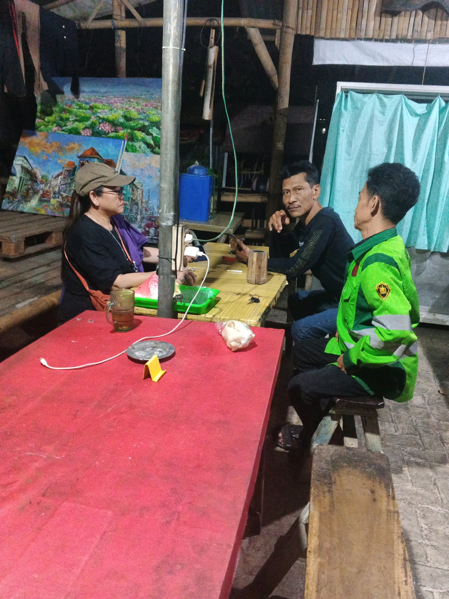 In the city of Ungaran, Indonesia, three individuals are seated around a table in an outdoor setting at night. A woman wearing a cap and glasses is on the left, facing right. In the center, a man in a black long-sleeved shirt with text visible on the sleeve is looking towards the camera. On the right, a man in a bright green jacket with reflective stripes is facing away from the camera, looking at the man in the black shirt. They are seated at a low, light-colored wooden table where there are drinks and snacks. A bright red table occupies the foreground. Behind the individuals, artwork is displayed on the wall, and a turquoise curtain hangs. The environment appears to be a simple, open-air establishment, possibly a food stall or restaurant, with bamboo construction visible overhead. Electrical cords are visible, along with a water dispenser.