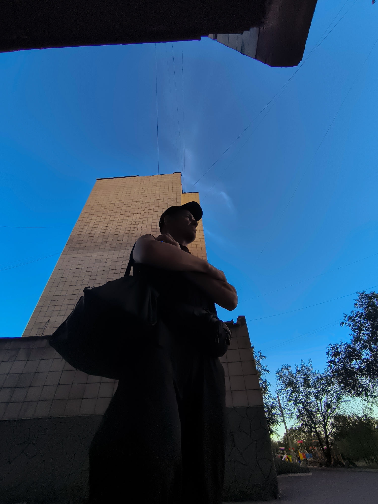 A low-angle, wide shot captures a silhouetted man wearing a baseball cap and dark clothing, with a duffel bag slung over his shoulder. He stands with his arms crossed, facing to the right of the frame. Behind him, a tall, beige, tiled building rises towards a clear blue sky. The top of the frame is dominated by a dark, overhanging structure, possibly a balcony or roof. To the right of the building, a playground with colorful equipment is visible amidst trees, suggesting an urban park or residential area. The scene appears to be set in the city of Qarağandı city administration, Kazakhstan.