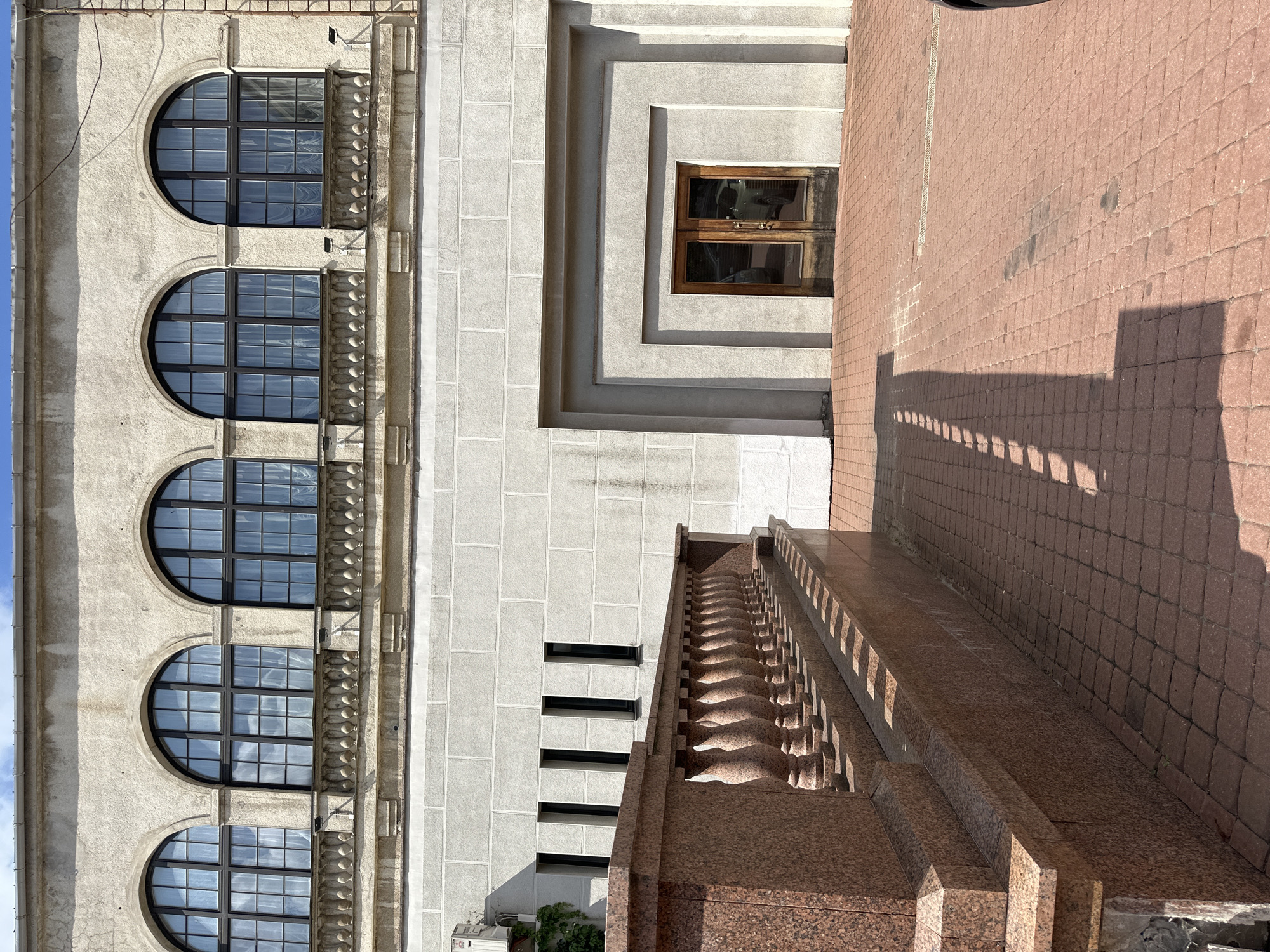 The image shows the exterior of a building in the city of Minsk, Belarus. The building has a facade with several arched windows on the upper level and a large entrance with double doors on the lower level. A red granite balustrade and pathway lead to the entrance. The overall architectural style appears to be classical with some modern elements. The sky is visible in the upper portion of the image, with some clouds present. The lighting suggests it is daytime, and the shadows indicate a sunny day. No people are visible in the image. The environment appears to be urban, with the building being the primary subject.