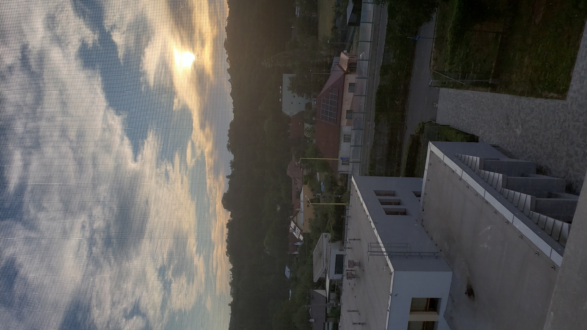 This is an eye-level, medium shot, outdoor photograph taken through a window screen, looking out over a suburban area of Prešov, Slovakia at sunset. The sun is visible in the upper right quadrant of the image, partially obscured by clouds and the window screen. The sky is filled with a mix of light and dark clouds, with warm tones of orange and yellow emanating from the sun.
In the middle ground, a residential area with several houses can be seen. One house in particular, located slightly to the left of center, has solar panels on its roof. The houses are nestled amongst lush green trees, and a hillside covered in trees forms the backdrop of the scene. There are a few streetlights visible, some yellow and some white.
In the foreground, the roofs of two buildings are visible. The building closest to the viewer is a light gray, modern-looking structure with a flat roof. It has an external ladder on its side. The second building, further in the foreground, also has a flat roof and appears to be part of a larger complex. A pathway made of gray paving stones leads away from the buildings into the green grass of a yard. The overall impression is one of a quiet, suburban evening in Prešov.