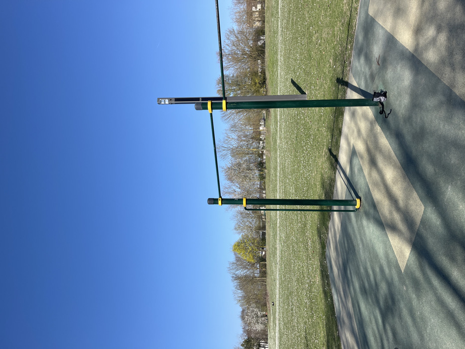 A green metal pull-up bar, with yellow accents, sits in a grassy area in the town of Saint-Brice-Courcelles, France. The bar is situated on a patch of artificial turf, a green and tan surface with a rectangular pattern. The ground is covered in patches of small white flowers. The bar is set against a backdrop of trees that are bare of leaves. The sky is a clear blue, with no clouds.