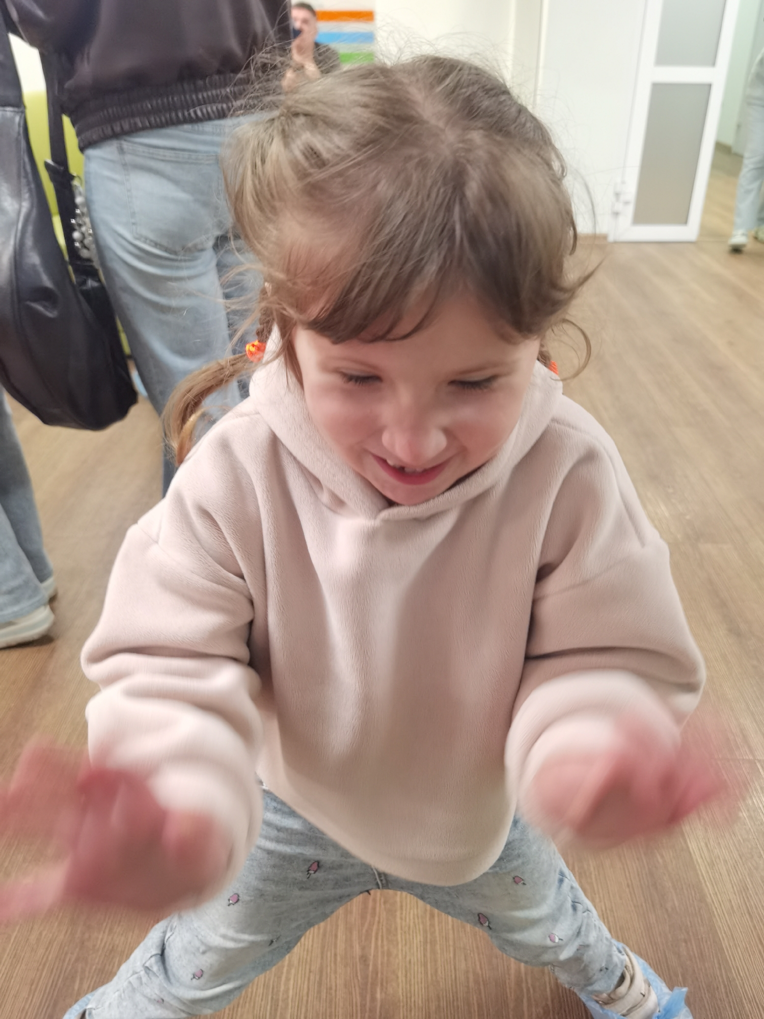 A young girl with light brown hair pulled back in pigtails, secured with orange hair ties, is positioned in the foreground, bending forward slightly. She is wearing a light pink, oversized hooded sweatshirt made of a soft, fleece-like material, and light blue jeans adorned with small, light pink ice cream cone patterns. Her hands are blurred due to motion, suggesting movement or clapping. She has a wide smile, looking downwards, indicating amusement or engagement with something below her.
Behind the girl, on the left, an adult's torso and legs are visible. This individual wears a dark bomber-style jacket and light blue denim jeans. A black shoulder bag with a beaded strap hangs from their shoulder. Further back and to the right, another adult is partially visible, wearing a dark top, with part of their face obscured by the first adult's hair. In the far background, a third person's lower legs and feet, wearing light blue shoe covers over white sneakers, can be seen near a light-colored door.
The setting appears to be an indoor space with light wooden flooring arranged in planks. The walls are white or light-colored, and a modern white door with a glass panel is visible on the right side of the frame.