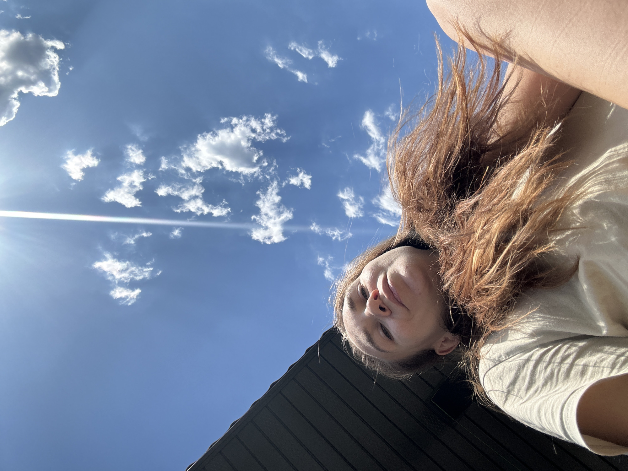 A low-angle, upward-facing photograph shows a woman with reddish-brown hair, wearing a light-colored t-shirt. The woman's face is prominent, with her nose pointing toward the camera. Behind the woman, the sky is blue with scattered white clouds. The sun is visible in the upper left portion of the frame. A dark, corrugated surface occupies the bottom portion of the image. The photo was taken outdoors, but there is no information to indicate the image was taken in Kharnavy, Russia.