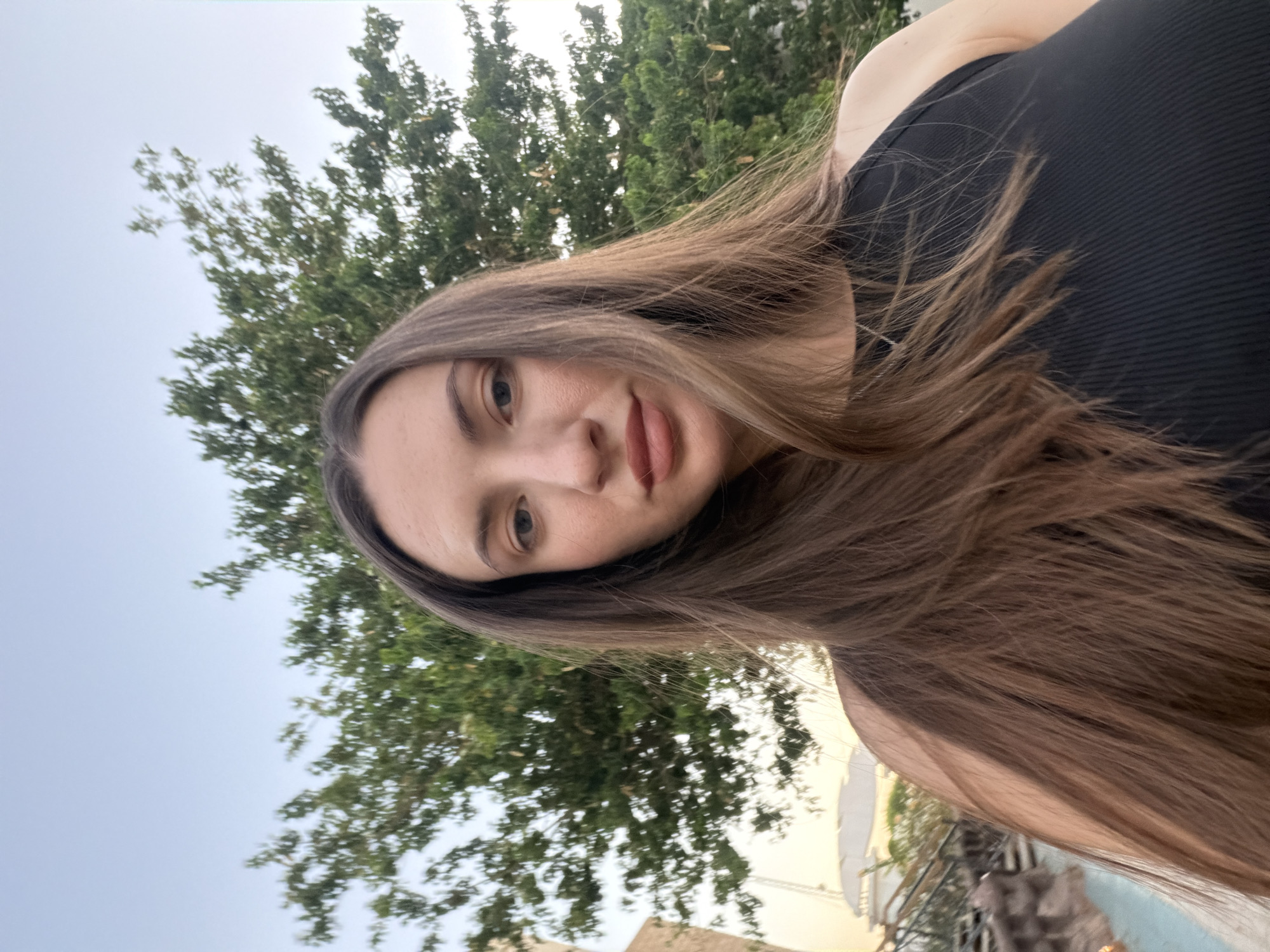 A young woman with long, dark brown hair is photographed from the chest up in an outdoor setting. She is wearing a black, ribbed tank top. Her head is tilted slightly to her left, and she is looking directly at the camera with a neutral expression. Her lips are painted with a matte, brownish-red lipstick. Behind her, there are green trees and a clear sky, suggesting a daytime scene. To her left, a portion of a building with architectural details and a glimpse of what appears to be a swimming pool area are visible, hinting at a resort or residential location. The lighting suggests it may be late afternoon or early evening. This scene is likely located in Sharm El Sheikh, Egypt, given the characteristic architecture and outdoor elements.