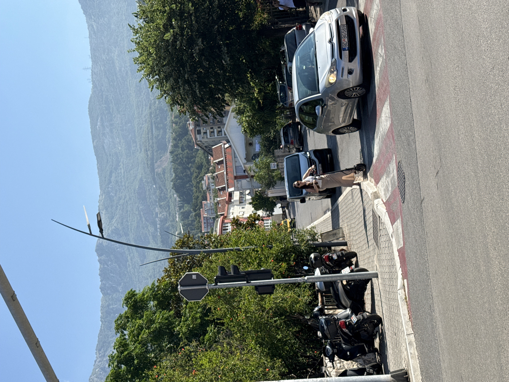 In Budva, Montenegro, on a sunny day, a woman on an electric scooter approaches a pedestrian crossing on a street lined with parked motorcycles. She is wearing a light-colored skirt and top. A silver Citroën C3, with license plate BD BG569, is stopped at the pedestrian crossing. Beyond the street, buildings with red-tiled roofs are visible, nestled against a backdrop of lush green hills and a hazy mountain range under a clear blue sky. Traffic lights and a stop sign are present at the intersection.