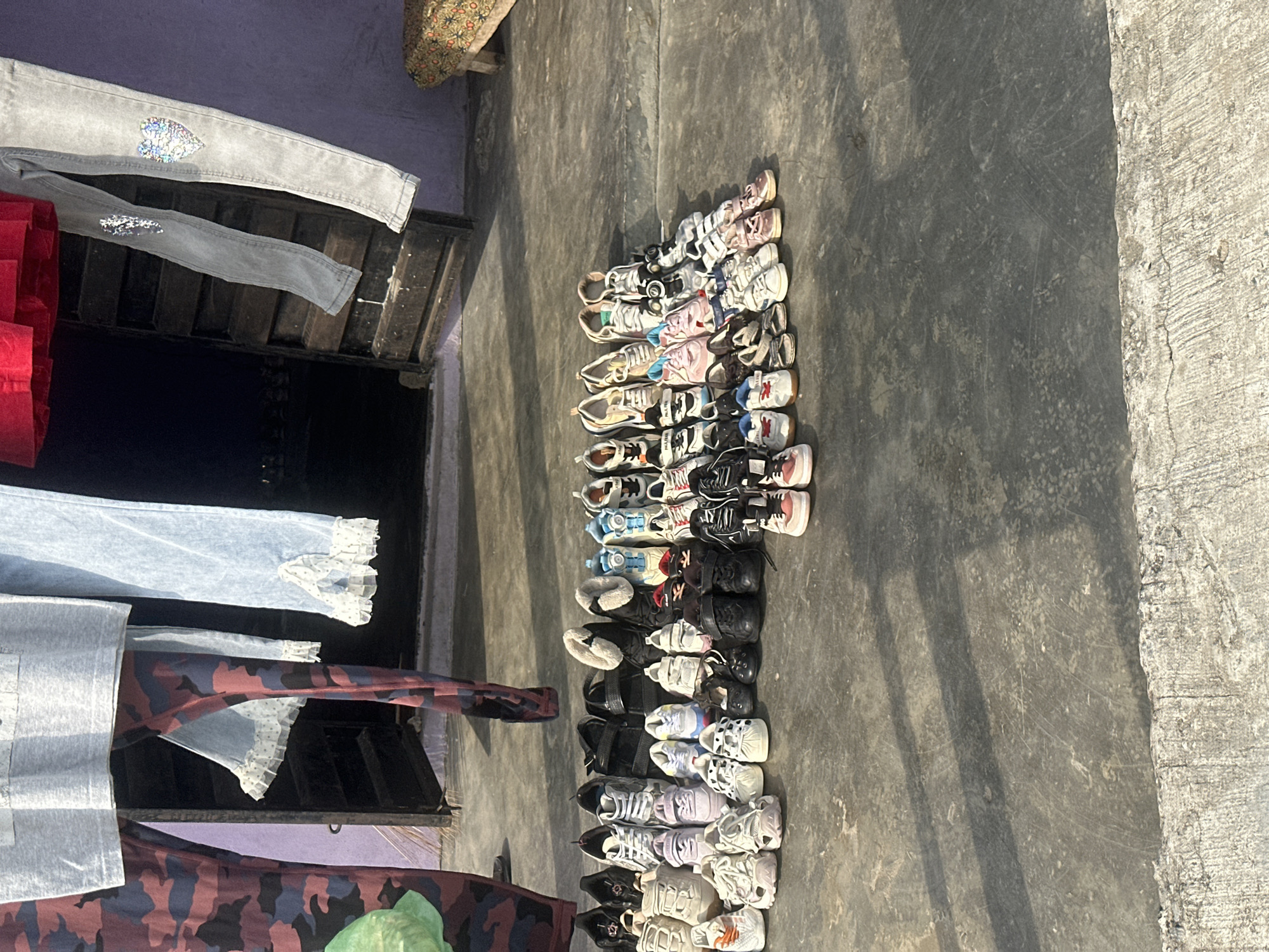 In Badagry, Nigeria, a collection of children's footwear is displayed on a concrete ground in front of a building. The shoes, primarily sneakers and sandals, are arranged in neat rows, suggesting they are for sale or distribution. The scene is lit by natural sunlight, casting shadows on the ground, and the background reveals a partially open doorway leading into a dark interior. Clothes hang on a line above, adding a domestic touch to the outdoor setting. The overall impression is one of everyday life in a Nigerian city, with a focus on the practical needs of its youngest residents.