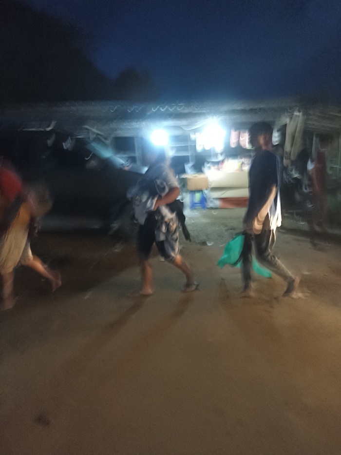 The image captures a blurred, low-light scene at night in Kecamatan Pujut, Indonesia, likely an outdoor market or street. Several people are visible, all with significant motion blur, indicating movement. In the foreground, three individuals are walking from left to right. On the far left, a person wearing a red top and light shorts is partially visible. A central figure, wearing a light-colored top and dark shorts, appears to be carrying an item in their arms. To their right, a third person in a dark long-sleeved shirt and light pants holds a light green object, possibly a bag.
The background features a simple market stall or shop, illuminated by several bright artificial lights that cast shadows from the walking figures onto the unpaved ground. Shelves, boxes, and various goods are faintly discernible within the brightly lit stall area. The sky above is dark, confirming the night-time setting. There is no visible text in the image. The scene portrays typical evening activity in a local commercial area.