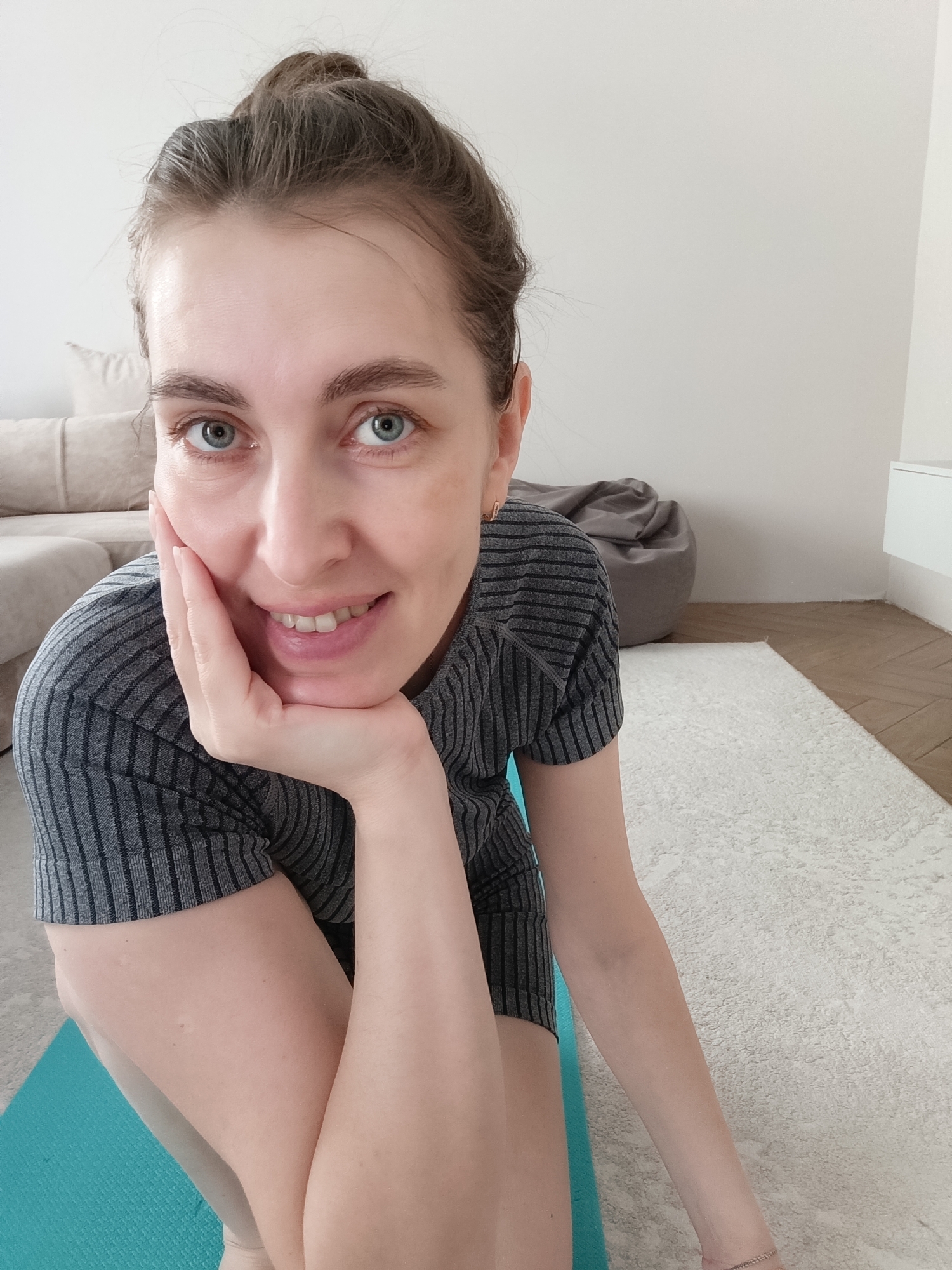 A woman with light skin and blue eyes is pictured in a close-up selfie. Her hair is pulled back into a bun. She is wearing a gray, striped, short-sleeved shirt. Her left hand is supporting her chin, and she is smiling at the camera. She is sitting on a teal yoga mat. The background includes a beige couch, a gray beanbag chair, a white wall, a white cabinet, a white carpet, and wooden flooring.
Based on the information available, I cannot confirm this image was taken in Chaikovsky, Russia. There is no information in the image itself that provides geographic location.