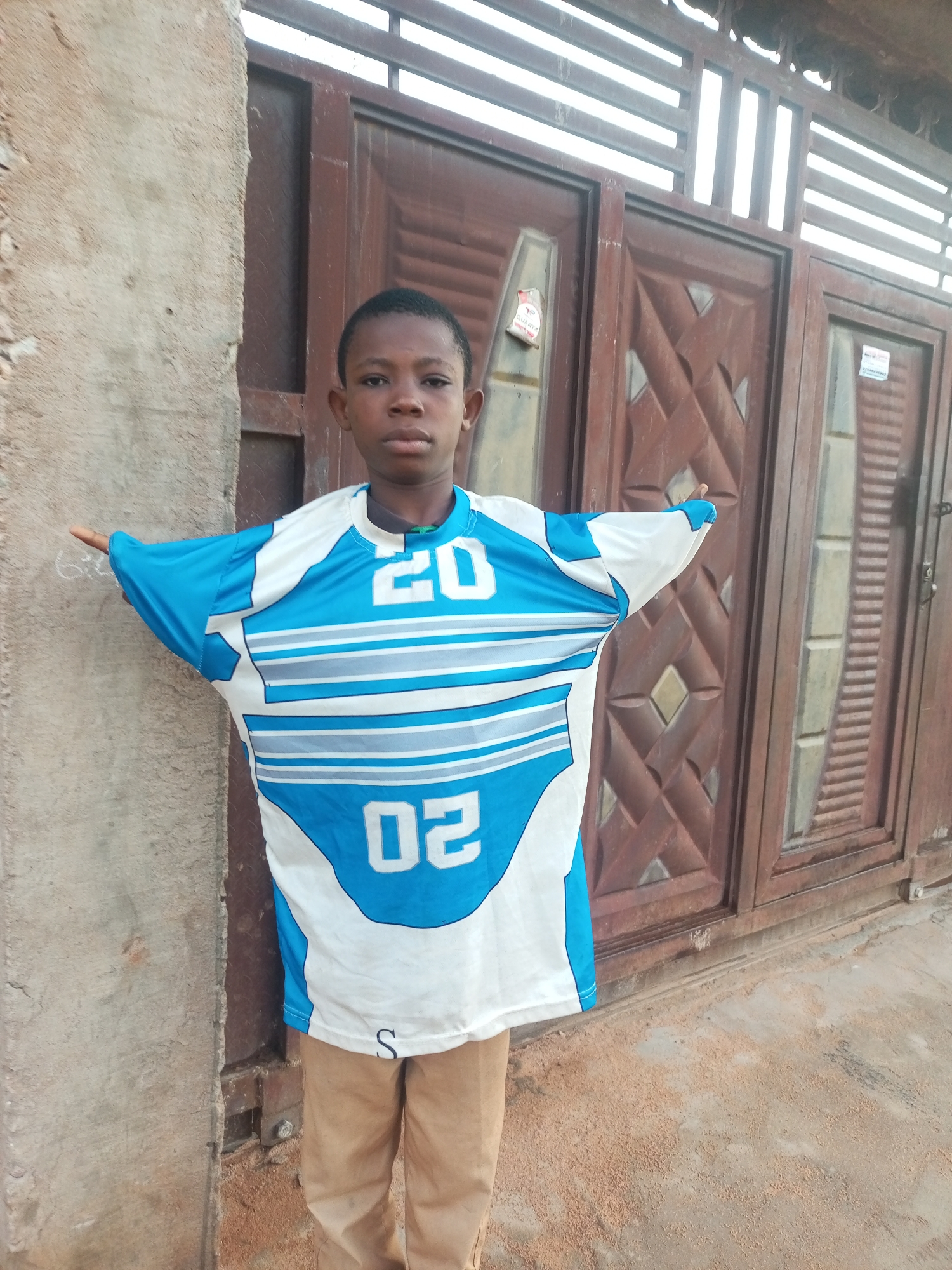 A young boy stands facing forward with his arms outstretched, wearing a blue and white t-shirt with the number "20" visible on the front, and the number "02" on the lower part of the shirt. He is also wearing tan pants. The boy is positioned in front of a brown, double door with a decorative pattern and a metal grate above it. To the left of the boy is a textured, light-colored wall. The ground in front of the door is covered with light brown dirt and debris. The scene appears to be outdoors, likely in a residential area. This image was taken in Tempure, Nigeria.