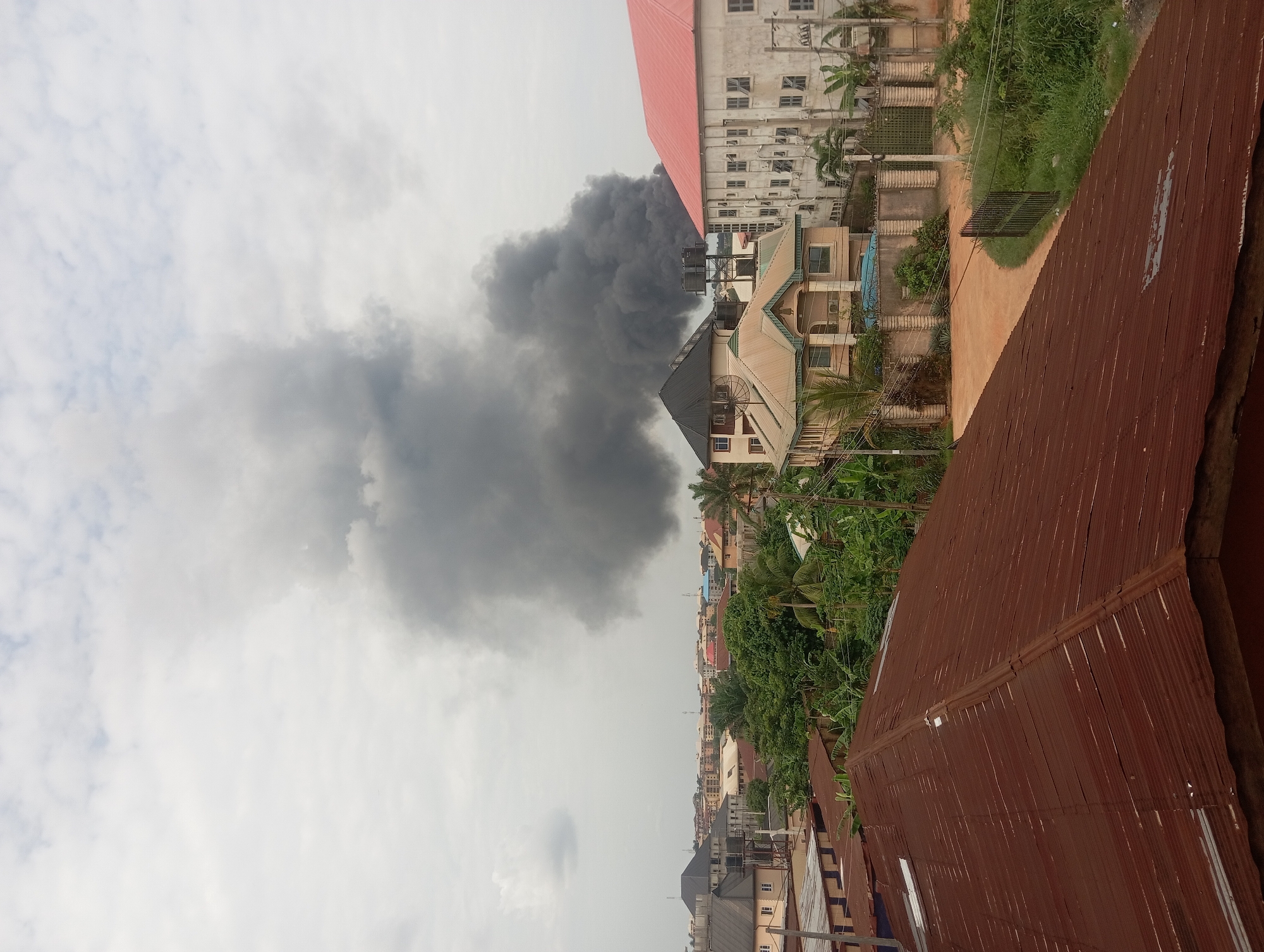 The image shows a large plume of dark smoke rising above the urban landscape of Nkpor, Nigeria. Buildings, including residences and a large structure with a red roof, are visible amidst lush greenery. The foreground features corrugated metal roofing. The sky is overcast with a mix of grey and white clouds.