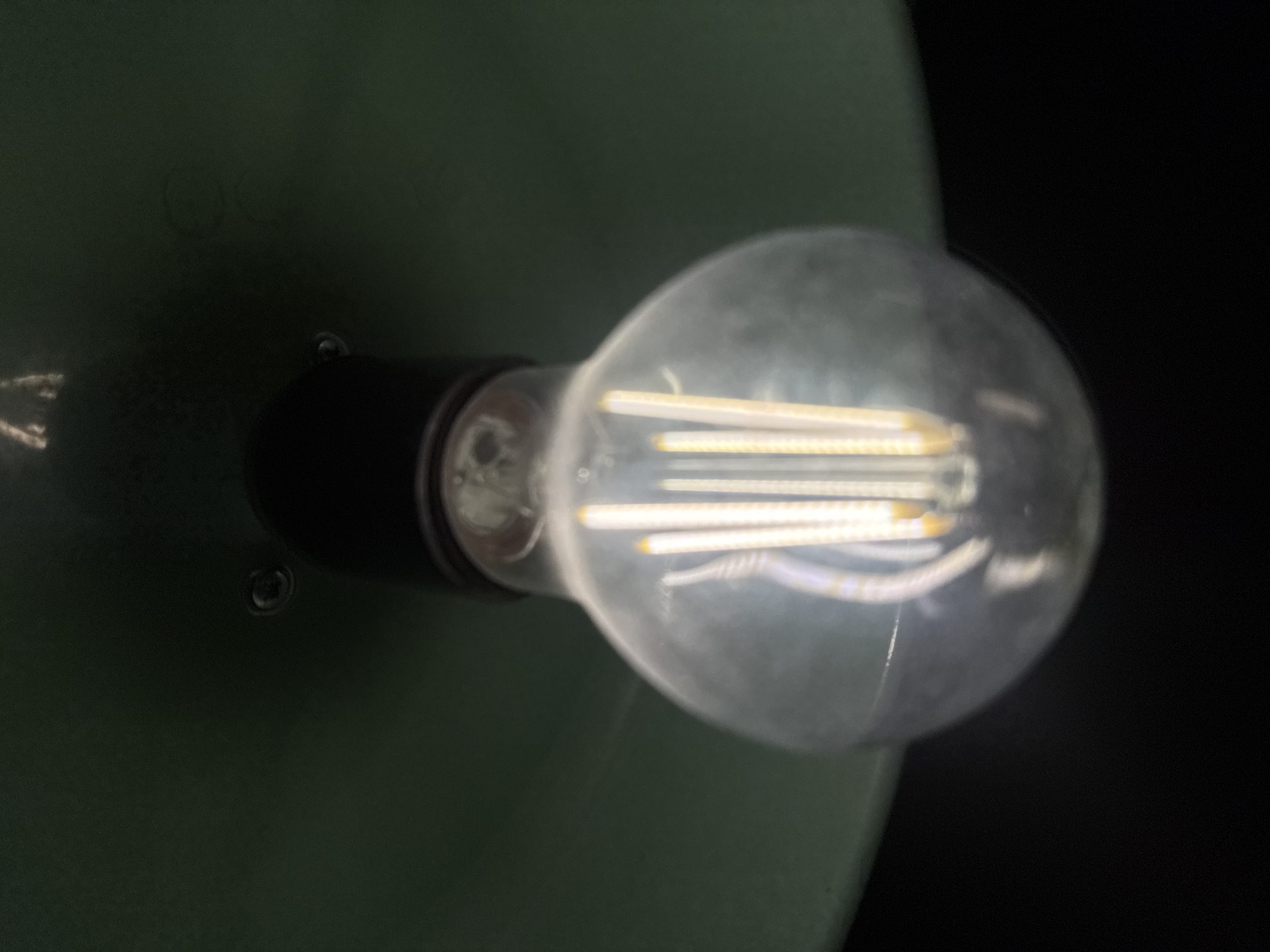 The scene is set in Yerevan, Armenia. The image captures a close-up, upward-facing view of an illuminated, clear glass LED light bulb. The bulb, which appears to be globe-shaped, features multiple vertical LED filaments emitting a warm, yellowish-white light. Some filaments exhibit a straight, strip-like appearance, while others present a coiled or bent configuration. The bulb is screwed into a dark, likely black, fixture base positioned immediately above it. Surrounding the base from above is a dark green, concave surface, consistent with the interior of a lampshade or housing. Two small, circular, reflective screw heads are visible on this dark green surface. The background below and to the sides of the bulb is uniformly black, indicating a dark environment where the bulb serves as the primary light source. No people or other active subjects are present.