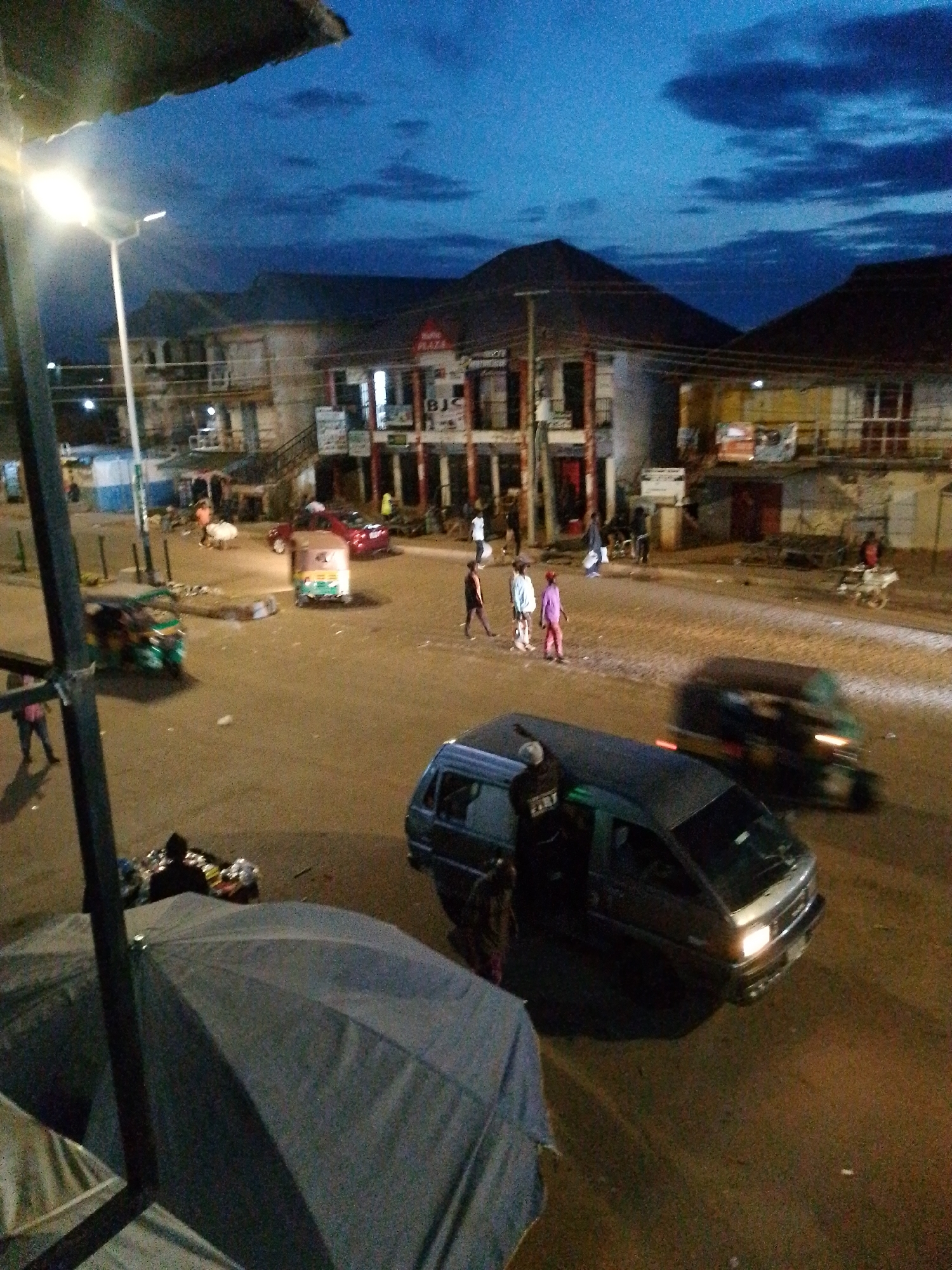 The image depicts a street scene in Bukuru, Nigeria, at dusk or early night. The sky is a deep blue with visible clouds, and artificial lights illuminate the street. In the foreground, a large, dark umbrella is partially visible, obscuring a portion of the lower left of the frame. Beyond the umbrella, a person in dark clothing is seated near a pile of items.
The street itself appears to be paved, with a few vehicles present. To the left, a green auto-rickshaw is parked, and further down the street, a darker auto-rickshaw is in motion, its lights illuminated. In the middle ground, a red car is also visible. Several individuals are walking along the street and sidewalks.
Buildings line the right side of the street, appearing to be shops or businesses, with some lit windows and signs. One sign reads "PLAZA". The architecture suggests a common urban style for the region. Poles and wires are visible overhead, indicative of street lighting and utility infrastructure. The overall impression is of a busy street at the end of the day.