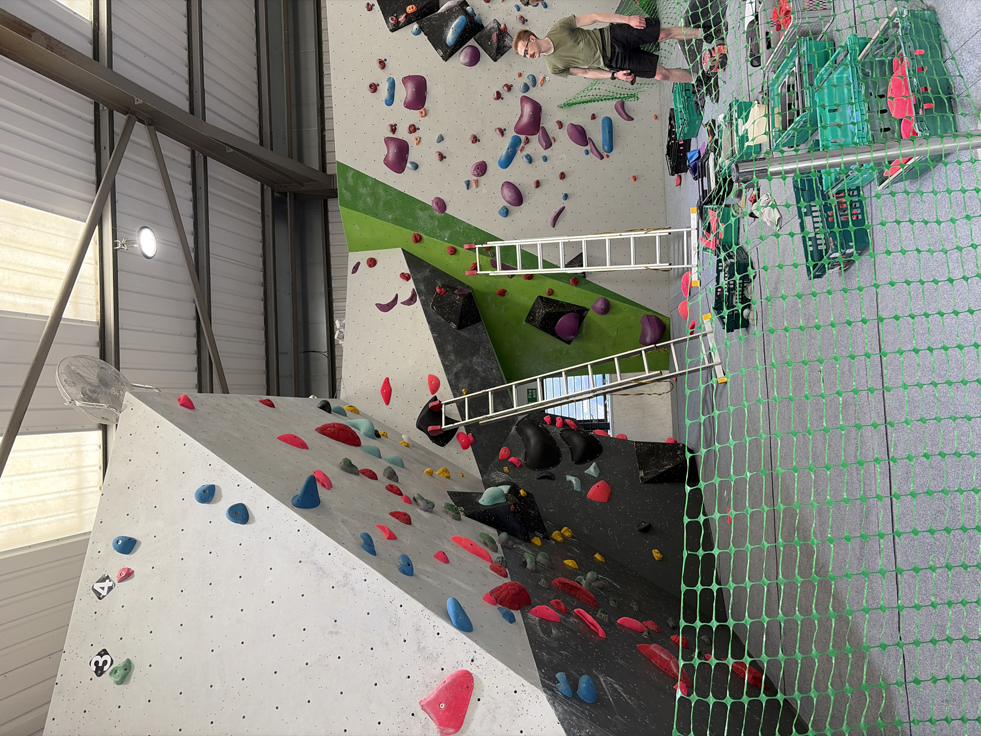 The image shows an indoor climbing gym in Ipswich, United Kingdom. The gym features a variety of climbing walls with different angles and holds of various shapes and colors. A man is standing on top of the wall. There is a ladder leaning against the wall. A green safety net partially obscures the view of the adjacent wall. Equipment containers and climbing gear are located to the right of the safety net. The gym's ceiling is visible, with metal beams and a ventilation system.