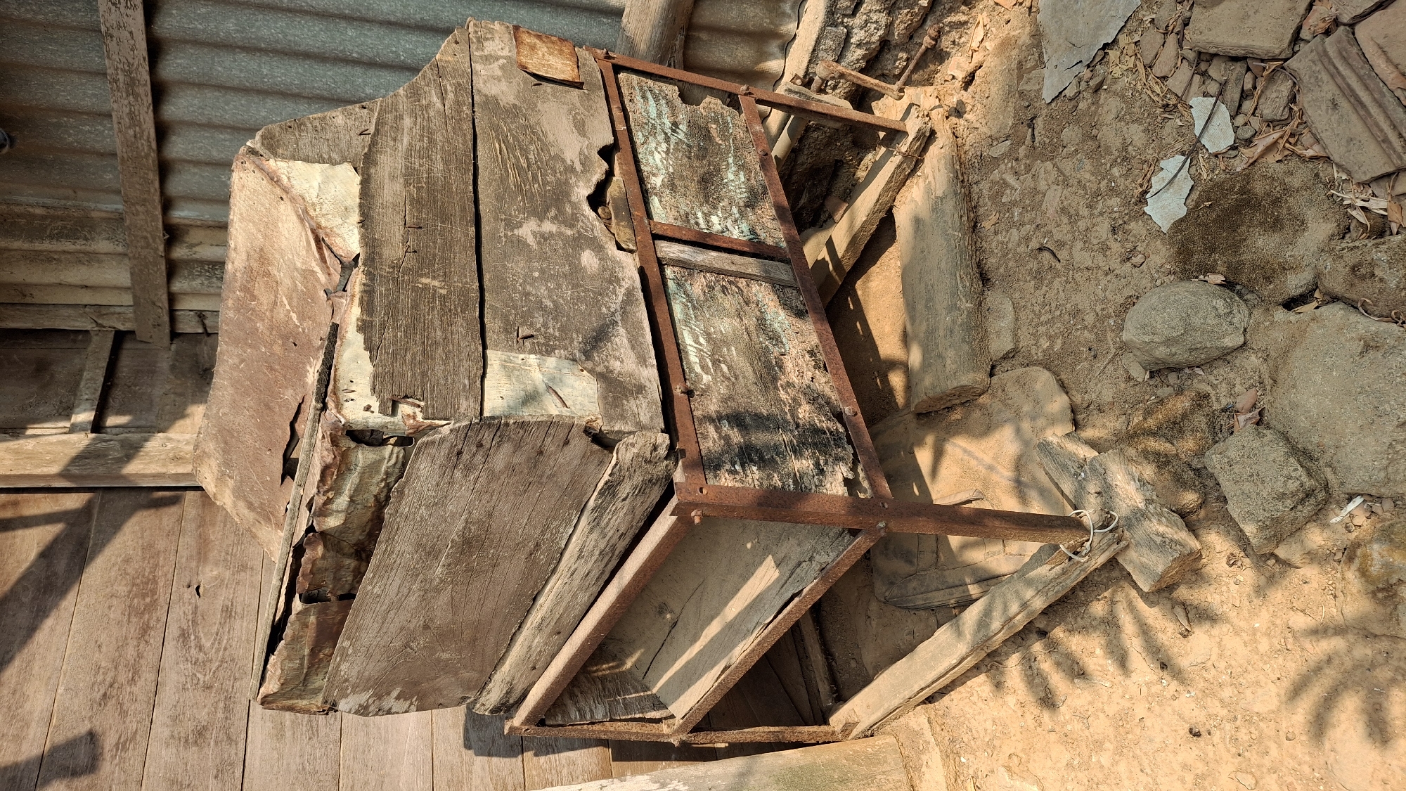 The image depicts an outdoor scene in Pucakwangi, Indonesia, showing a weathered, abandoned structure. A large, rustic wooden box with a rusty metal lid is precariously stacked on top of a metal-framed shelf unit. The shelf unit has two compartments, each with dilapidated wooden fronts, revealing glimpses of what appears to be stored material or empty space within. The structure is set against a backdrop of weathered wooden planks and corrugated metal siding, suggesting it is part of an old building or shed. The ground surrounding the structure is uneven and consists of dry soil, scattered stones, and debris, including broken pieces of pottery or tiles. Sunlight casts distinct shadows from foliage onto the ground and the structure, indicating a daytime setting. The overall impression is one of disuse and decay, with the elements having taken their toll on the materials. There are no people or animals visible in the image, and no apparent activity is taking place.