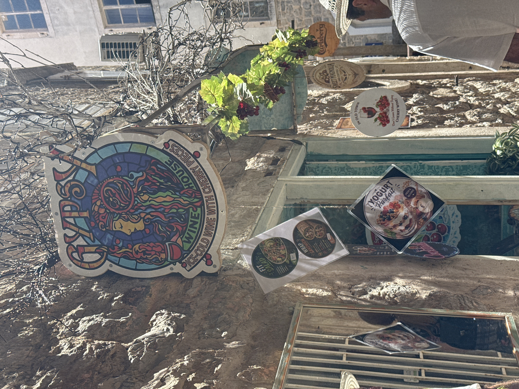 The image captures a sunlit street scene in the Old Town of Budva, Montenegro, focusing on the entrance of a commercial establishment named "Gypsy Wines House." A prominent, colorful, stained-glass-style circular sign for "GYPSY WINES HOUSE ICE-COLD WINE WITH CHERRY PIECES" hangs centrally, depicting a woman with elaborate headwear and a goblet. Above it, bare branches, possibly adorned with string lights, intertwine. Below the main sign and to the left, several square promotional menus are suspended, displaying items such as "FIT SALAD," "WRAP SANDWICH," and "YOGURT Parfait."
To the right of the main sign, artificial green grapevines with red clusters hang as decoration. Further down on the right, additional circular and oval signs are visible: a white one stating "IN OLD TOWN Don't Leave Without Trying CHERRY WINE!," a brown "Sweet Magnolia AND COFFEE" sign, and an orange "Carlsberg" sign. The building walls are constructed from rough, light-colored stone. On the far left, a metal gridded door or window guard is partially visible. In the background on the right, an older building with windows and an external air conditioning unit is present.
A man wearing a straw hat and a white short-sleeved shirt is seen from behind on the far right, walking away from the camera. The scene is brightly lit by natural sunlight, casting distinct shadows and highlighting the textures of the stone walls and various signs.