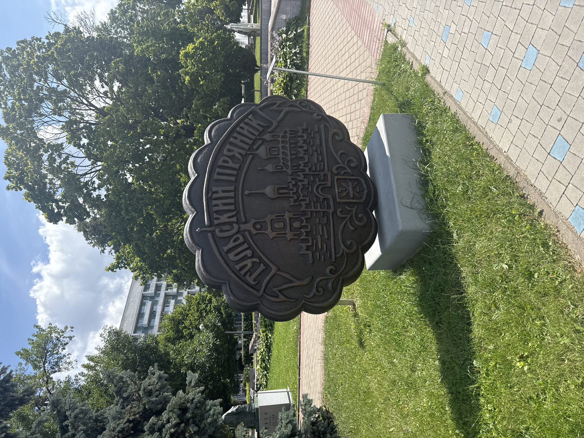 A circular, relief sculpture with a scalloped edge and a darker, metallic finish is displayed in a park setting. The sculpture depicts a cityscape with architectural elements and Cyrillic text at the top. The text reads "ТУЛЬСКІЙ ПРЯНИК" which translates to "Tula Pryanik" (a type of gingerbread). The sculpture is mounted on a light grey rectangular concrete base.
The sculpture is positioned in a grassy area. To the left of the sculpture, a dark green evergreen tree is partially visible. Behind the sculpture, lush green grass and manicured bushes are present. Further in the background, a light-colored, multi-story building with many windows can be seen, surrounded by mature green trees. A paved walkway with a patterned surface of light grey and blue tiles runs along the right side of the sculpture. The sky above is blue with scattered white clouds.
The sculpture is a monument to the Tula Pryanik, a famous type of gingerbread from Tula, Russia. It is located in Mahilyow, Belarus.
