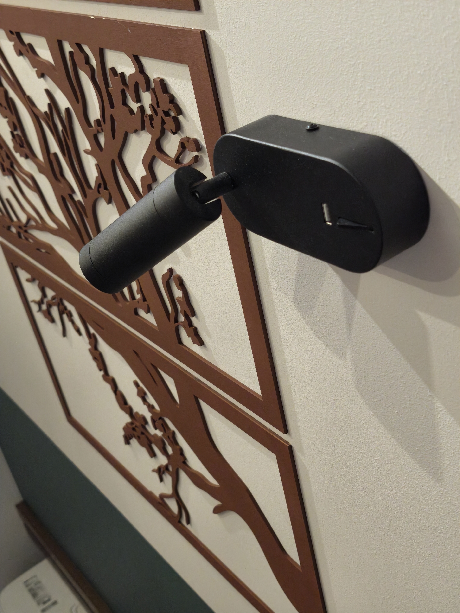 A close-up shot shows a black, wall-mounted spotlight positioned next to a decorative wall panel featuring a stylized, brown-toned tree motif. The spotlight, with its cylindrical head and oval base, is the primary focus, affixed to a textured, off-white wall. The decorative panel, located slightly behind the spotlight, adds depth to the composition with its intricate, laser-cut tree design. The scene is indoors.