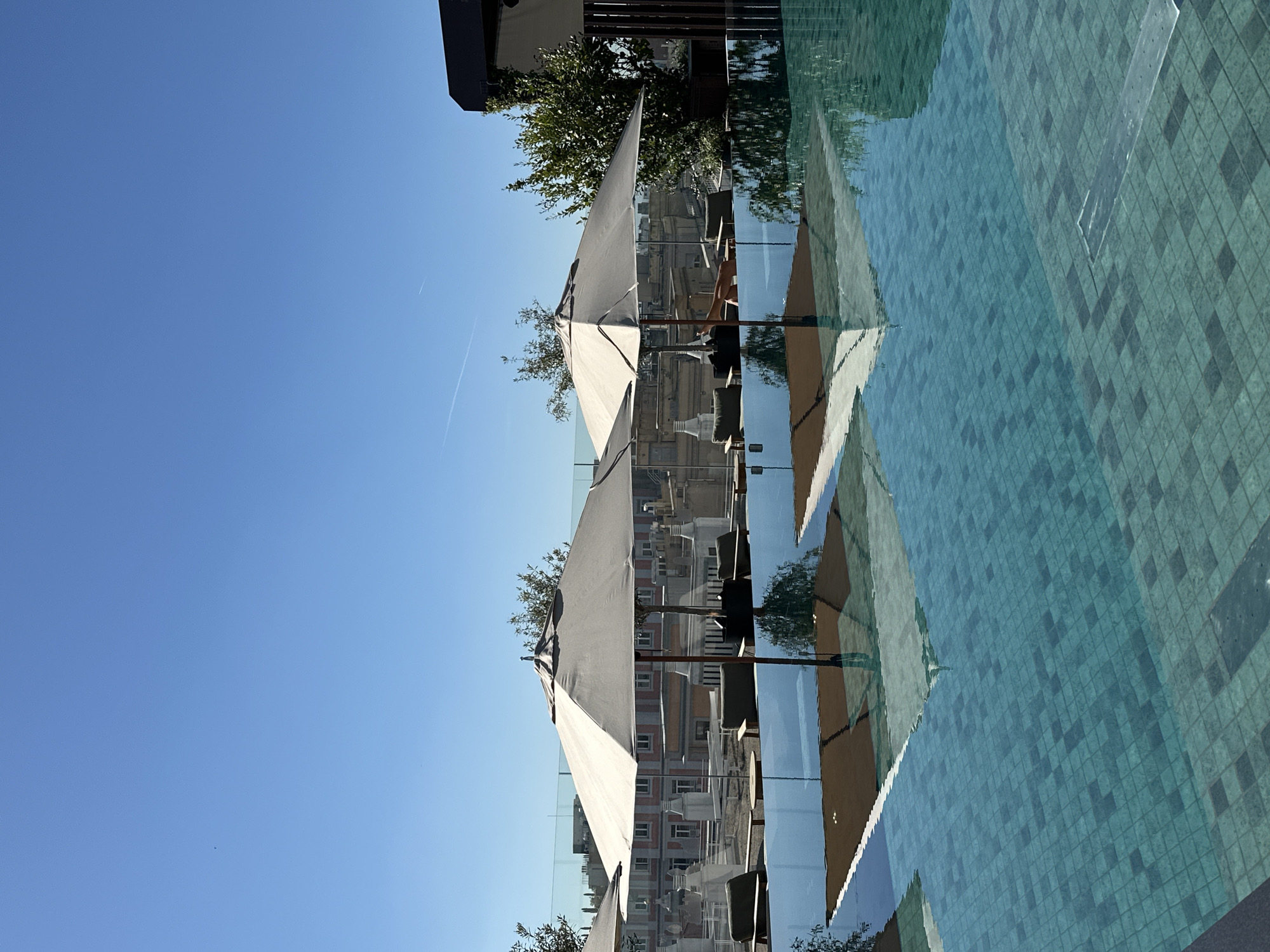 The image shows a view from a rooftop pool in Madrid, Spain, featuring several white umbrellas casting reflections on the water's surface. In the background, buildings typical of Madrid's architecture are visible through a glass railing. The sky is clear blue with a visible contrail. No people are readily visible, but there is a hint of chairs and tables suggesting a seating area. The pool's tiling is aqua-colored with darker accents.