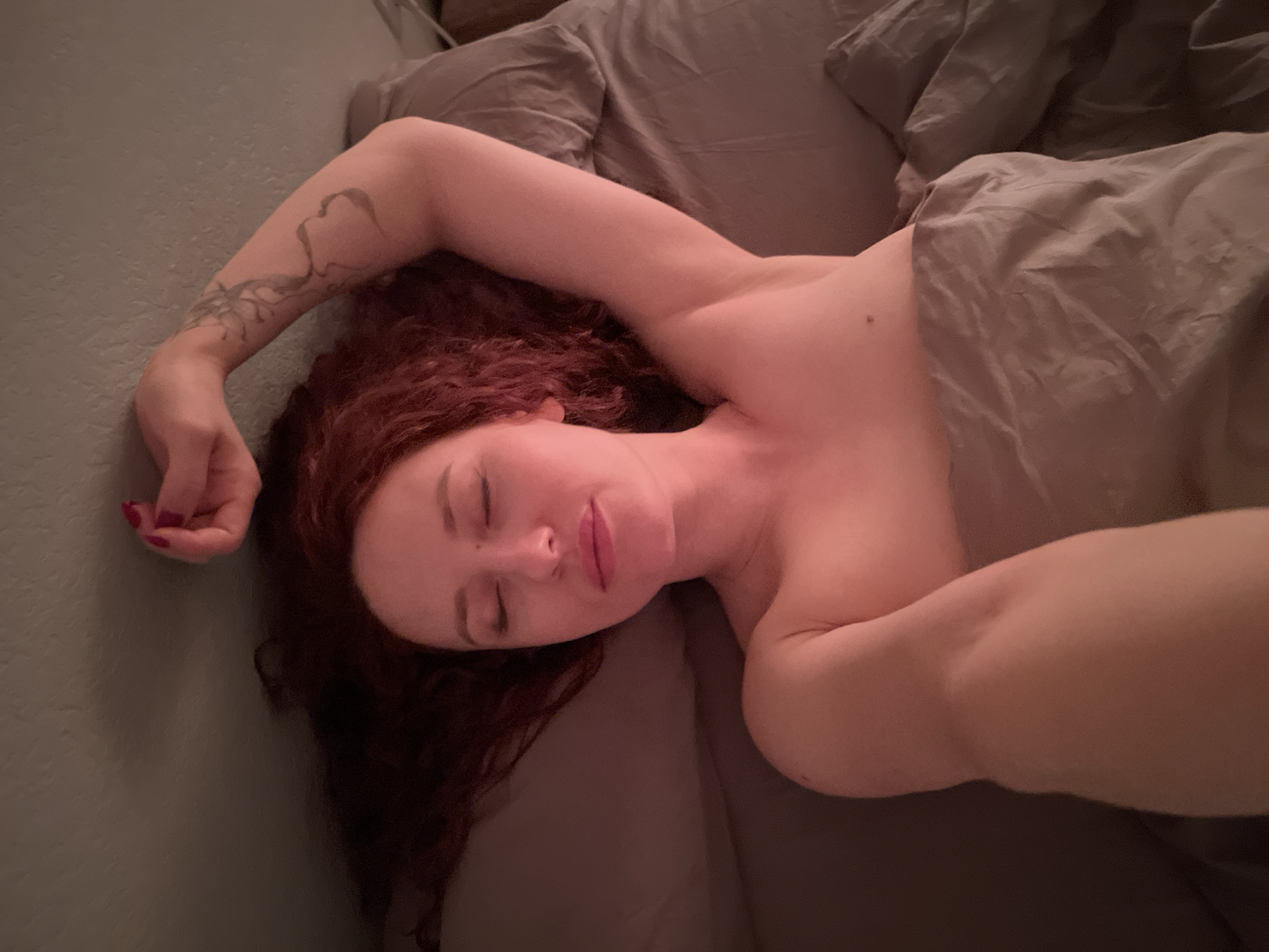 A fair-skinned person with long, curly red hair is lying in a bed with gray sheets and comforter. The person appears to be asleep, with their eyes closed. One arm is extended above their head, and the other arm is outstretched beside their body. A tattoo is visible on the extended arm. The background includes a light gray wall. There is no indication of the location being Saint Petersburg, Russia.