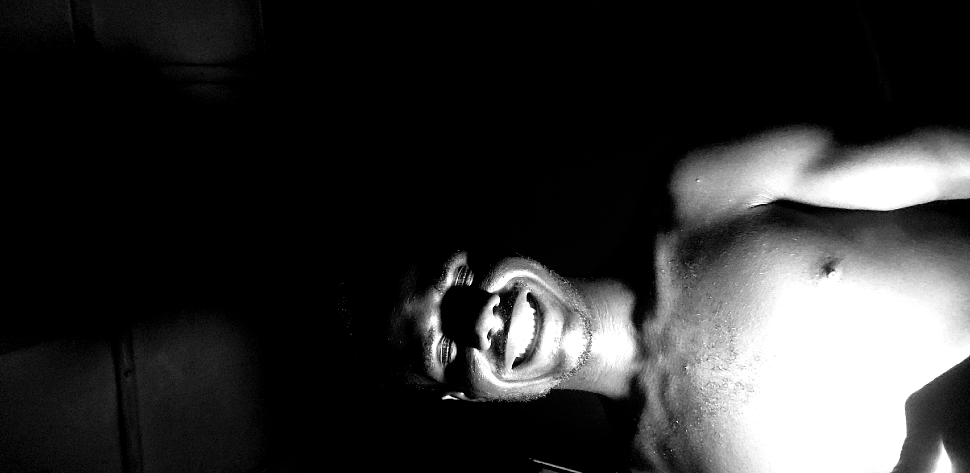 A black and white, close-up, low-angle, selfie-style shot captures a shirtless Black man smiling broadly in a dimly lit indoor environment, possibly in Narayi, Nigeria. The man's face is brightly illuminated from below, highlighting his teeth, lips, and the contours of his face and neck. He has short hair and a light beard. His chest and shoulders are visible, glistening with what appears to be sweat or water droplets. The background is almost entirely dark, with a sliver of what might be a ceiling or upper wall visible at the top of the frame. The lighting creates a dramatic and intense effect, with strong shadows obscuring details of the surroundings.
